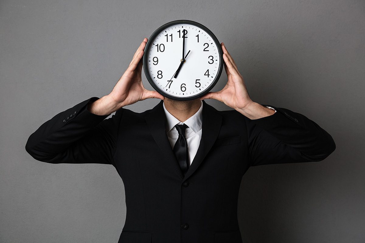 A man in a suit holding an analogue clock up to cover his face, the time reads 7:00