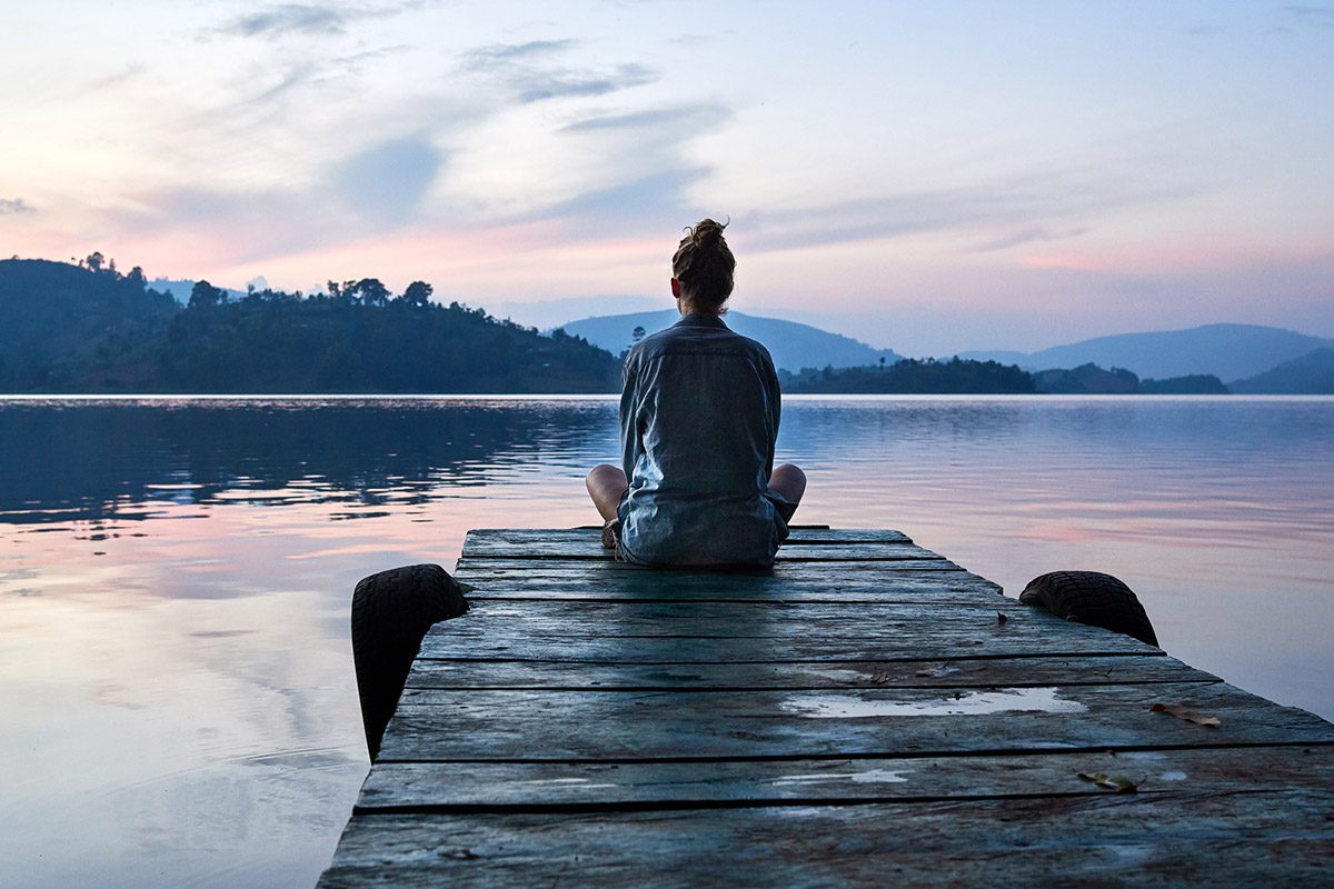 A woman sitting cross legged at the end of a dock looking out at a lake.