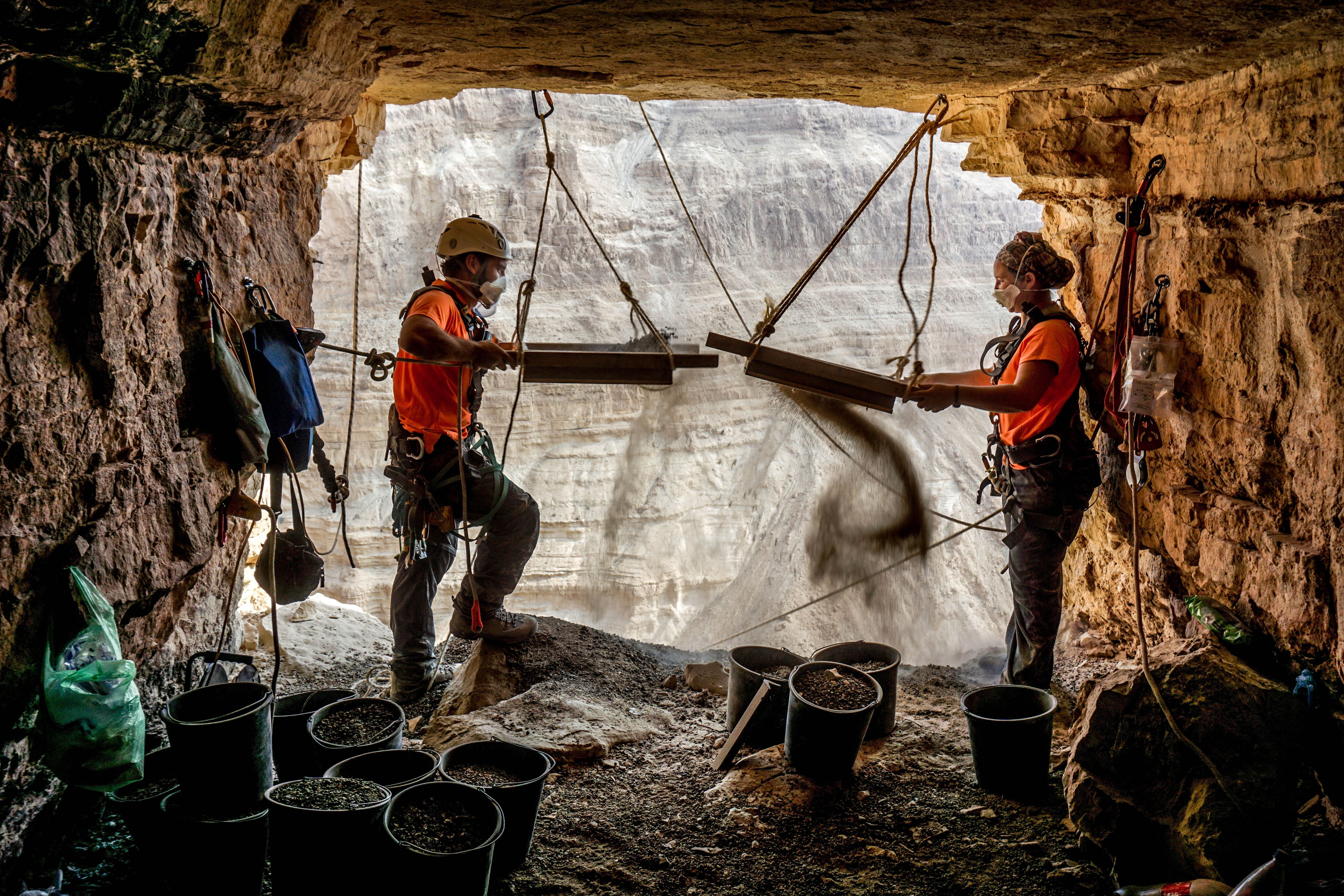 Two scientists in a cave they have rappelled into holding up seives where they are sifting the dirt for ancient finds Two scientists in a cave they have rappelled into holding up seives where they are sifting the dirt for ancient finds