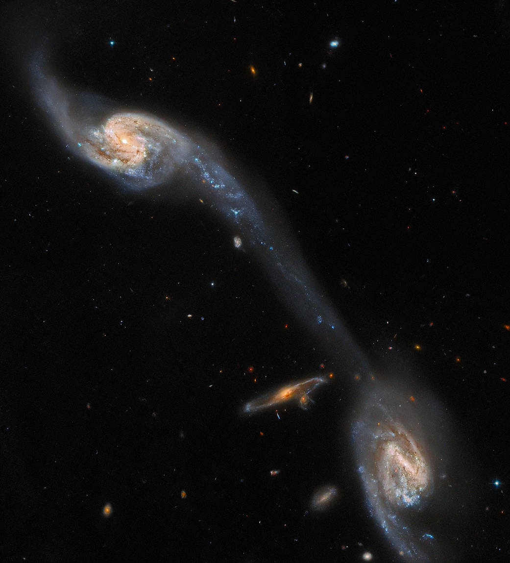 Two spiral galaxies are viewed almost face-on; they are a mix of pale blue and yellow in colour, crossed by strands of dark red dust. They lie in the upper-left and lower-right corners. A long, faint streak of pale blue joins them, extending from an arm of one galaxy and crossing the field diagonally. A small spiral galaxy, orange in colour, is visible edge-on, left of the lower galaxy.