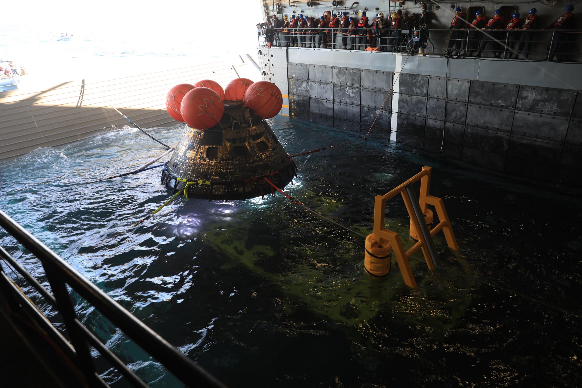 The craft is in water within the walls of the ship. People line a platform looking at the spacecraft.