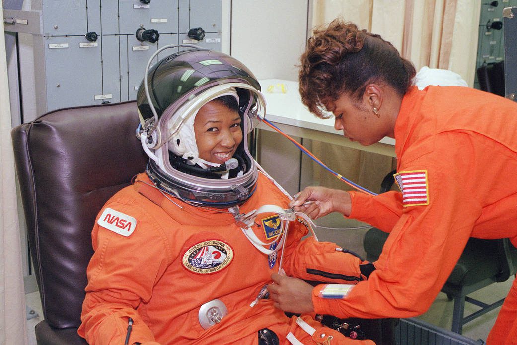 Astronaut Mae Jemison is staring at the camera while McDouglke is asjust a strap in the astronaut suit.