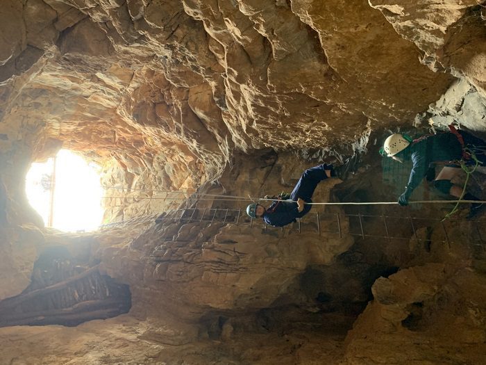 Cave entrance with bright sunlight and person hangs in a harness descending into the cave.
