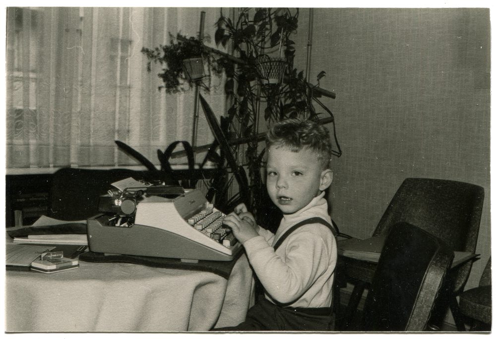 An antique photo of Baby Boomer boy in the 1960s sitting at a table with his hands on the keys of a typewriter