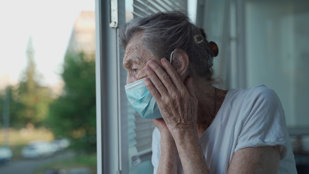 Old woman in medical mask looks worriedly out of window