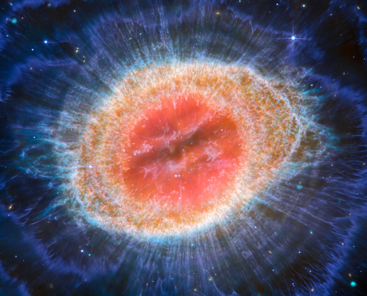 This image of the Ring Nebula appears as a distorted doughnut. The nebula’s inner cavity hosts shades of red and orange, while the detailed ring transitions through shades of yellow in the inner regions and blue/purple in the outer region.