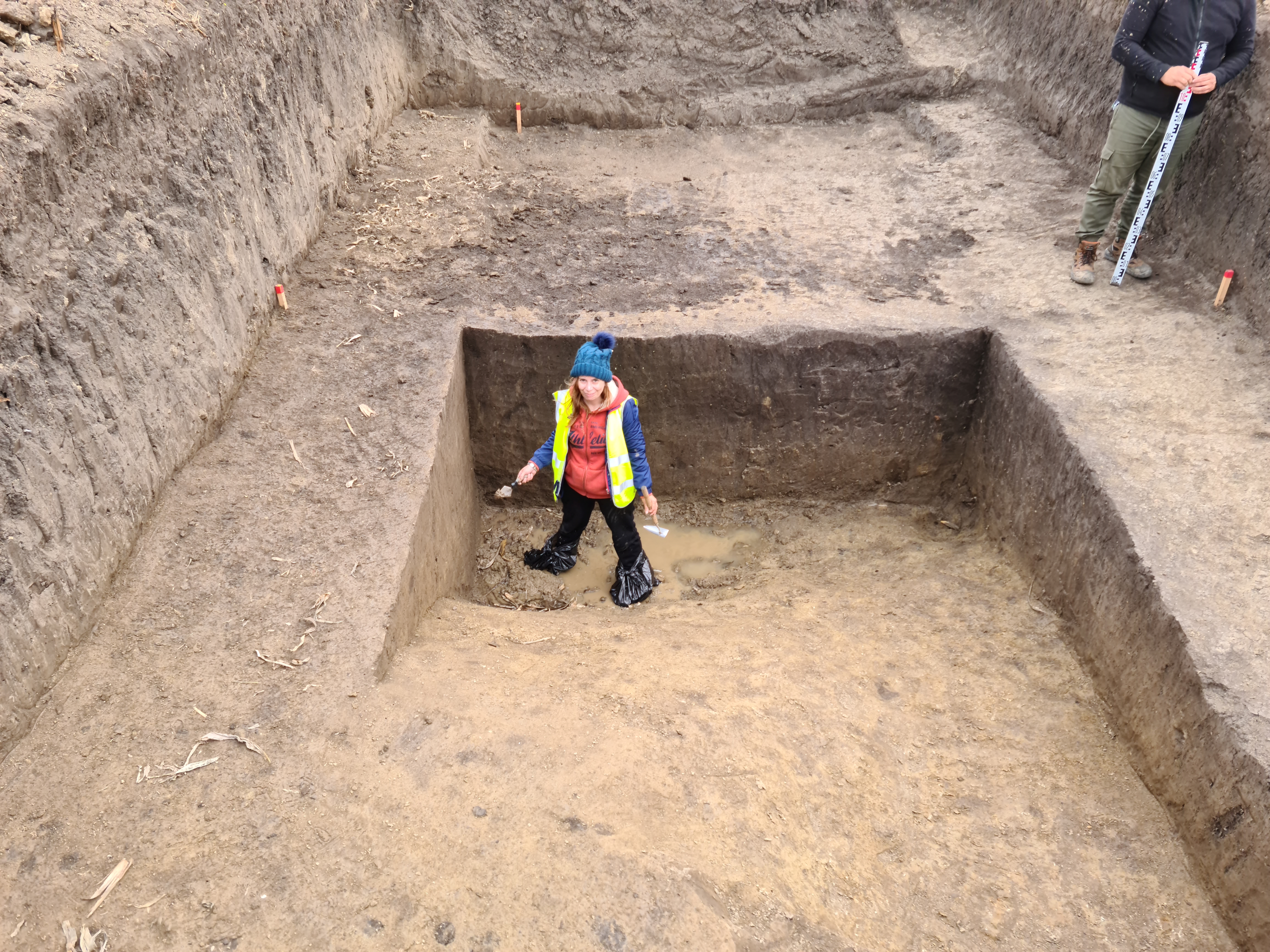 archaeologist wearing blue hat, red hoody and hi viz vest stands in deep hole at excavation site