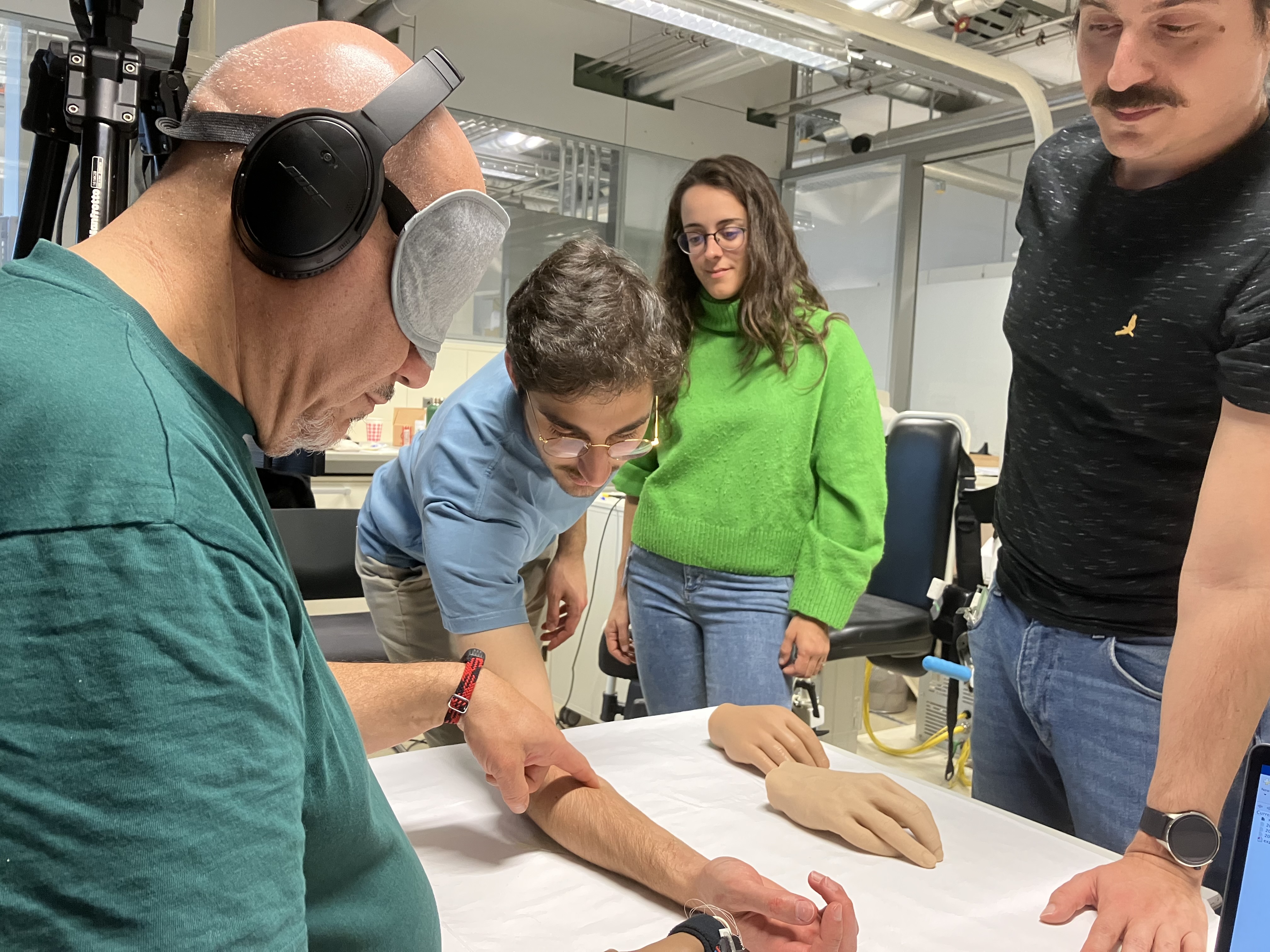 blindfolded man touches another man's arm using his prosthetic limb, there are other prosthetic hands on the table and a man and woman stand watching