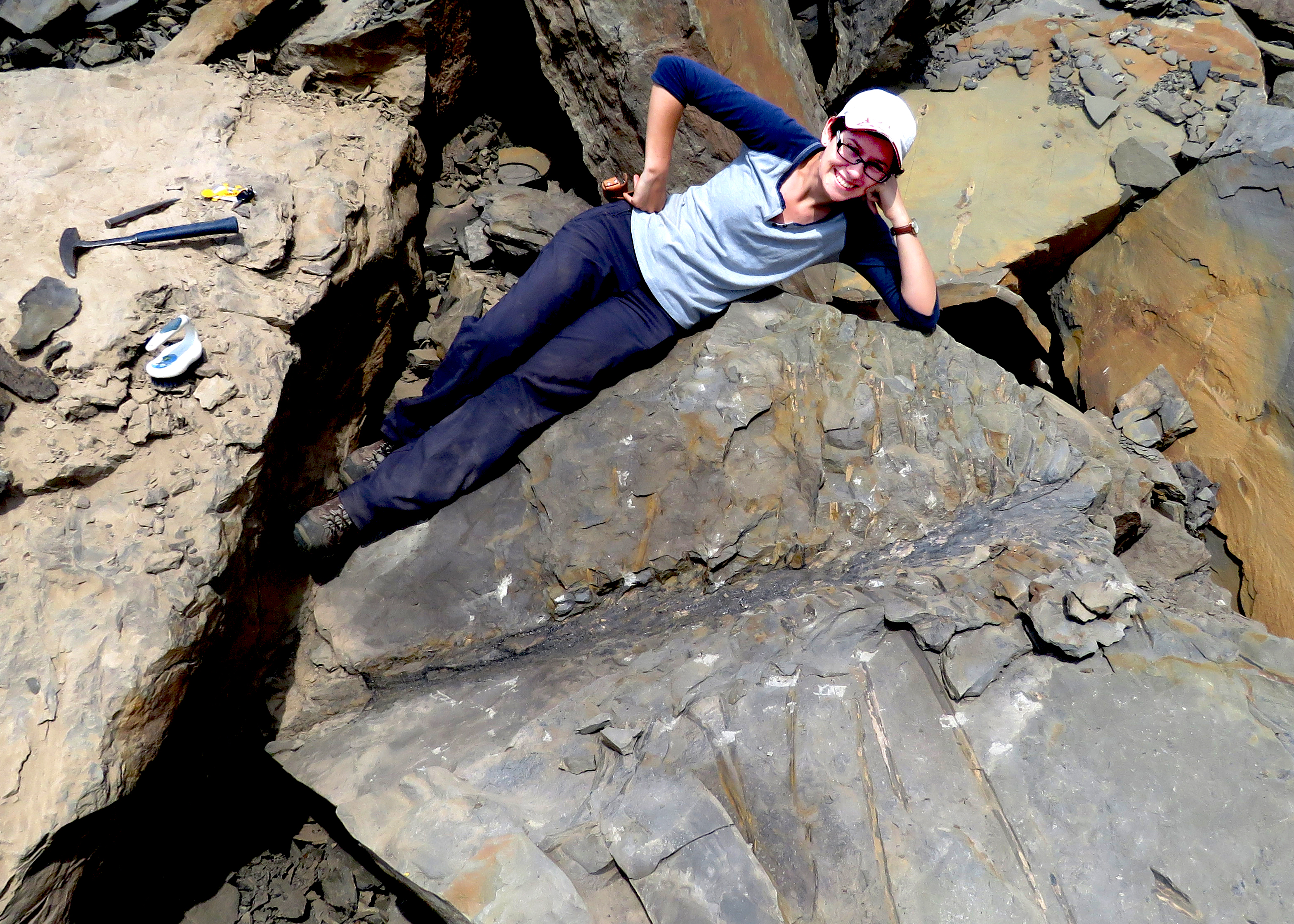 Researcher wearing blue trousers and blue shirt with cap lying on a rock above the Sanfordiacaulis fossil looking very happy. Small tools are near by.