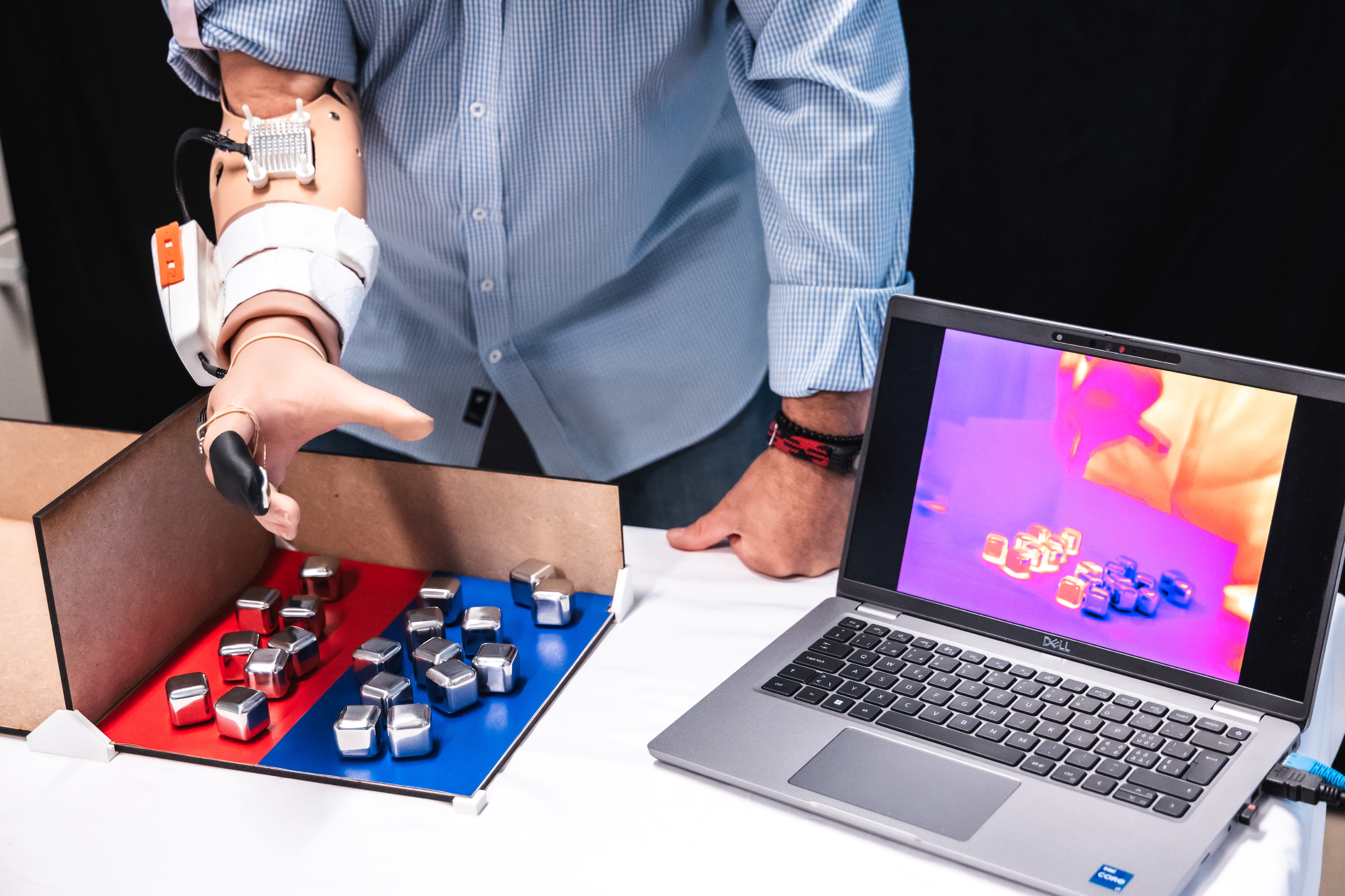 man with prosthetic arm sorting metal cubes into two piles, one on a red background and one on a blue background; laptop next to them on the table shows thermal imaging, with the red cubes hot and the blue cubes cold