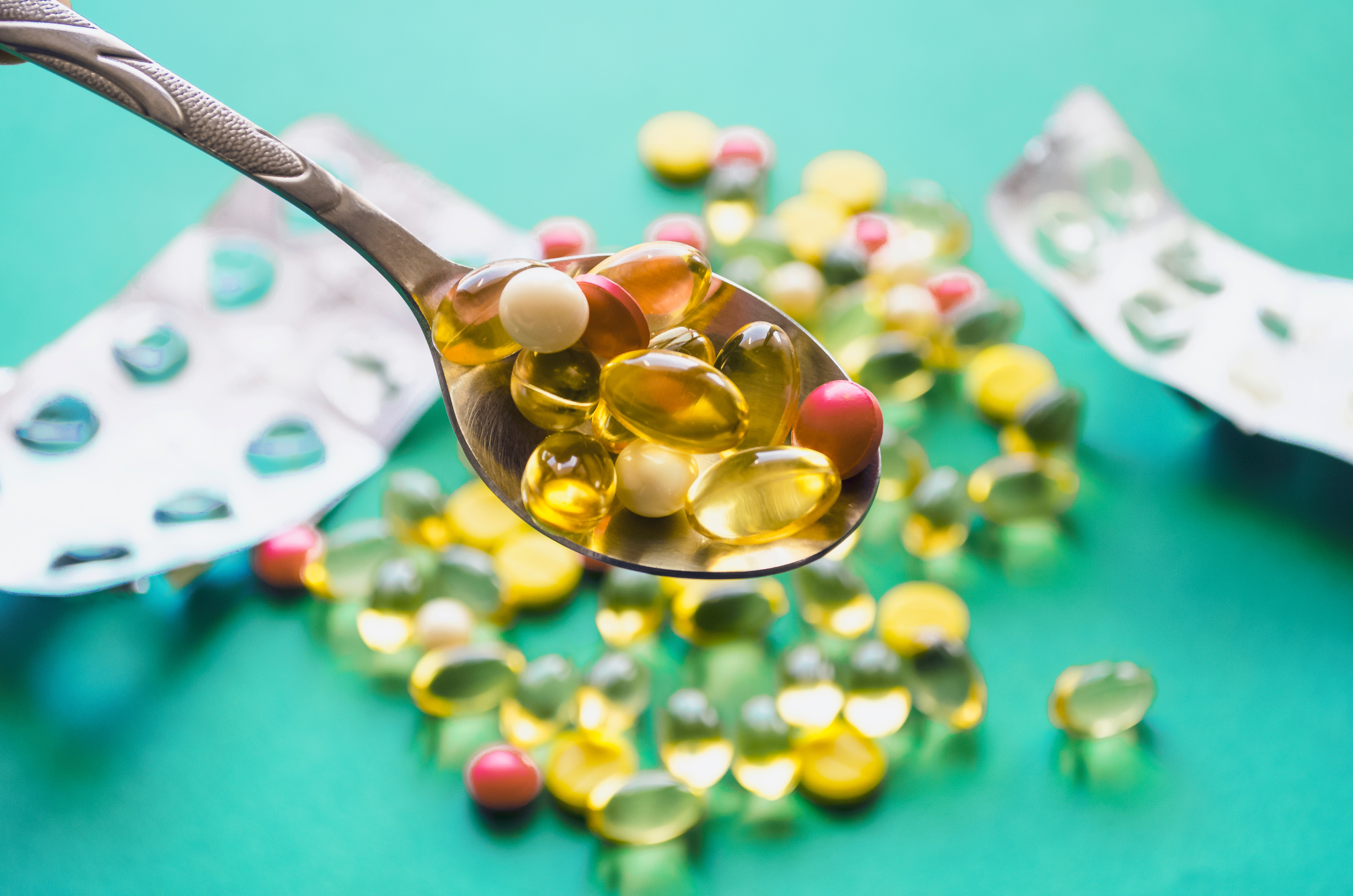 silver spoon full of different coloured capsules and tablets, with out of focus golden capsules and blister packets against turquoise background