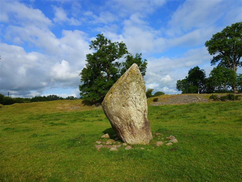 A close up photo of the standing stone at Mayburgh Henge, Cumbria, England. The stone rises to a point and has irregular sides. It is surrounded by smaller stones at its base. In the background you can see part of the henge mound that encloses the stone, along with several trees. 