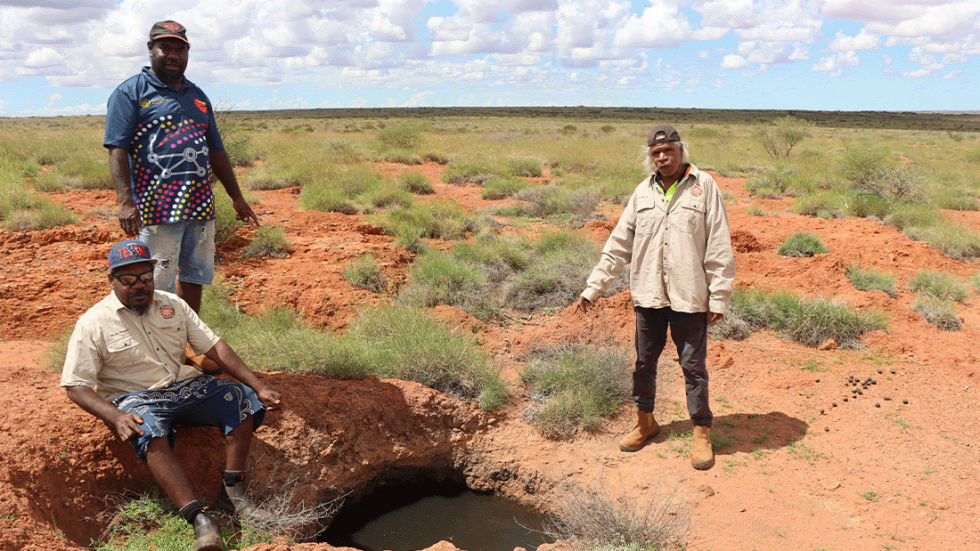 Three men stand in the desert near a small hole filled with water. These are the KJ Martu Rangers.