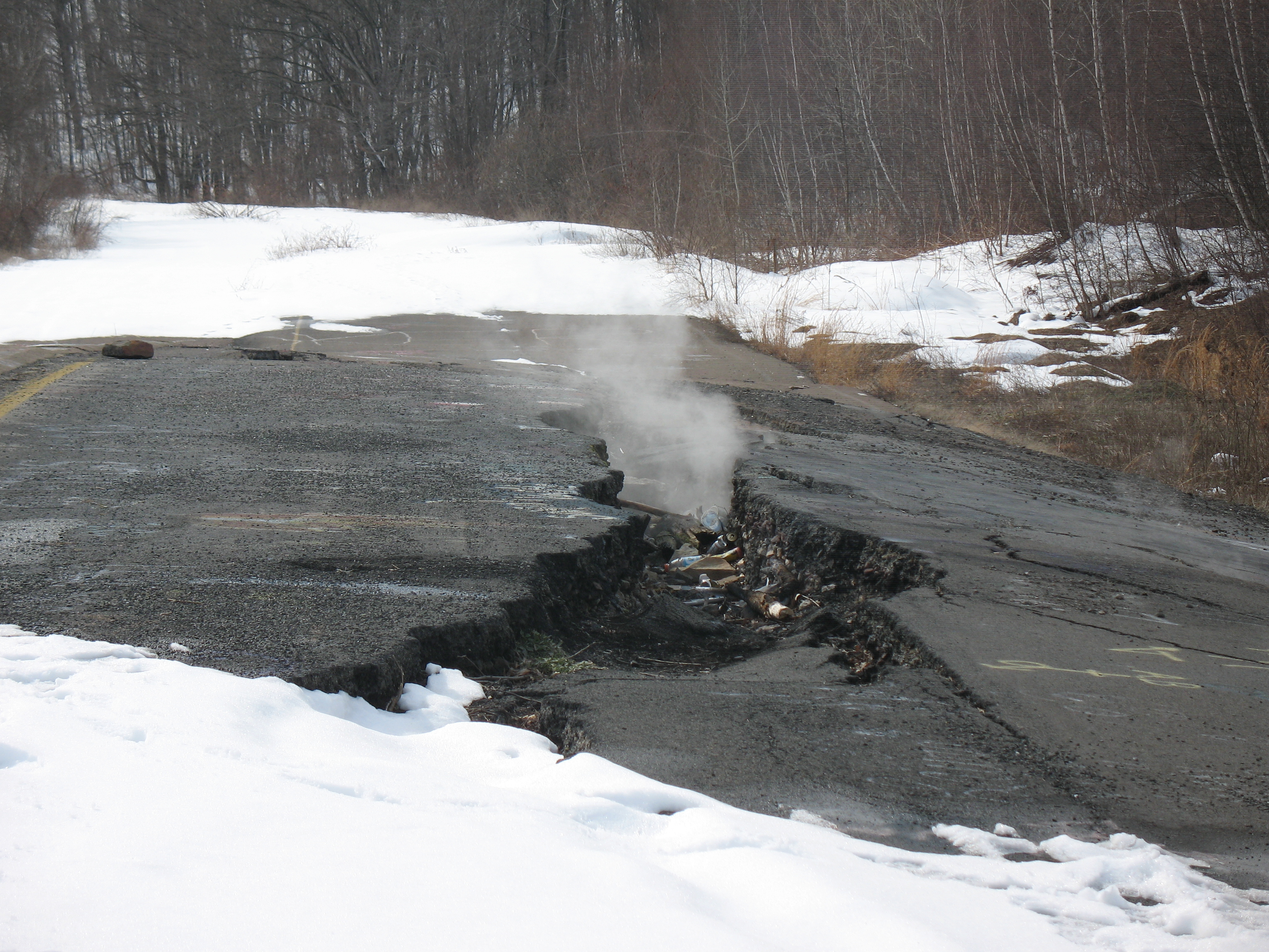 Plumes of smoke leak from a large fissure in the tarmac of an abandoned road in Centralia. The road it surrounded by snow and spars trees but there is a large patch of ground in the middle of the photo that is clear due to the heat. 