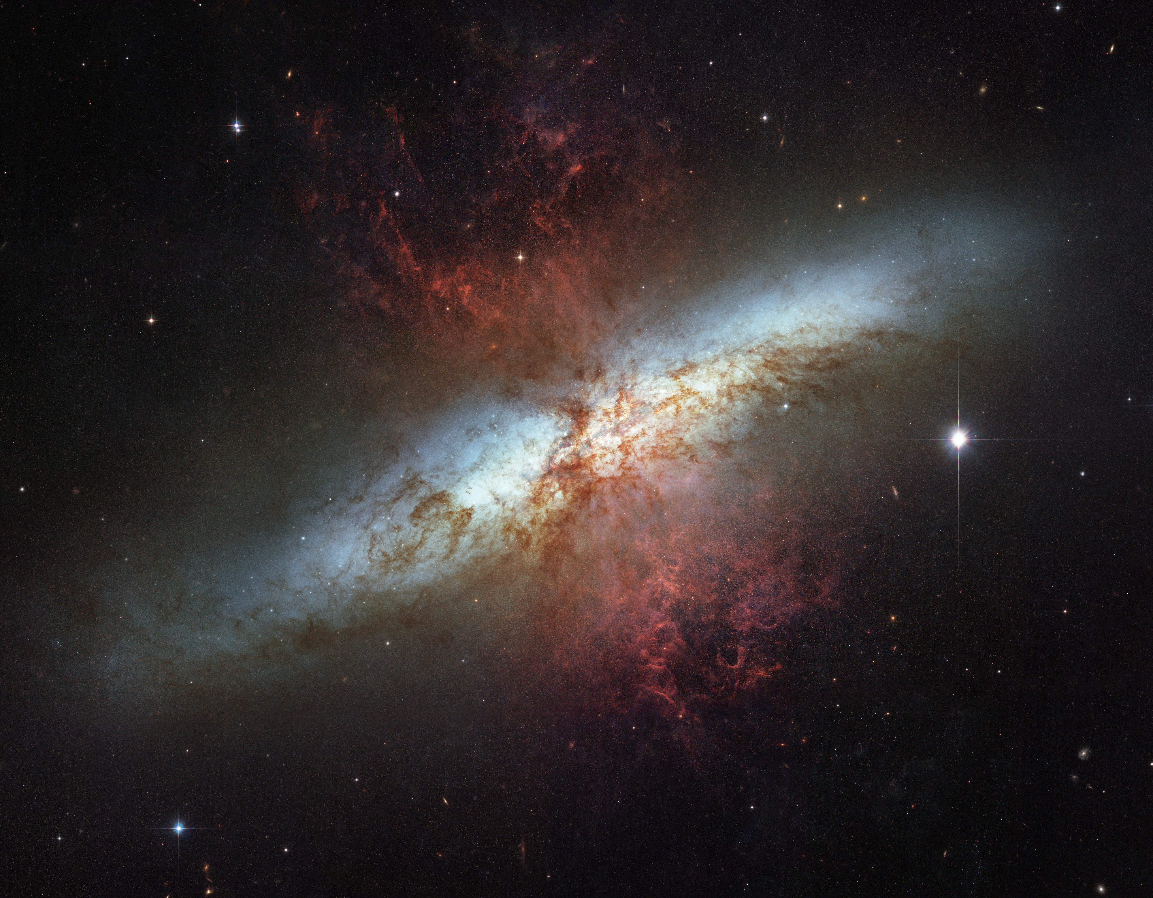 The galaxy is messy. its main body is a bright blue-white fuzzy region. On top of it at ninety degrees an hourshape glass of red dust and gas is visible