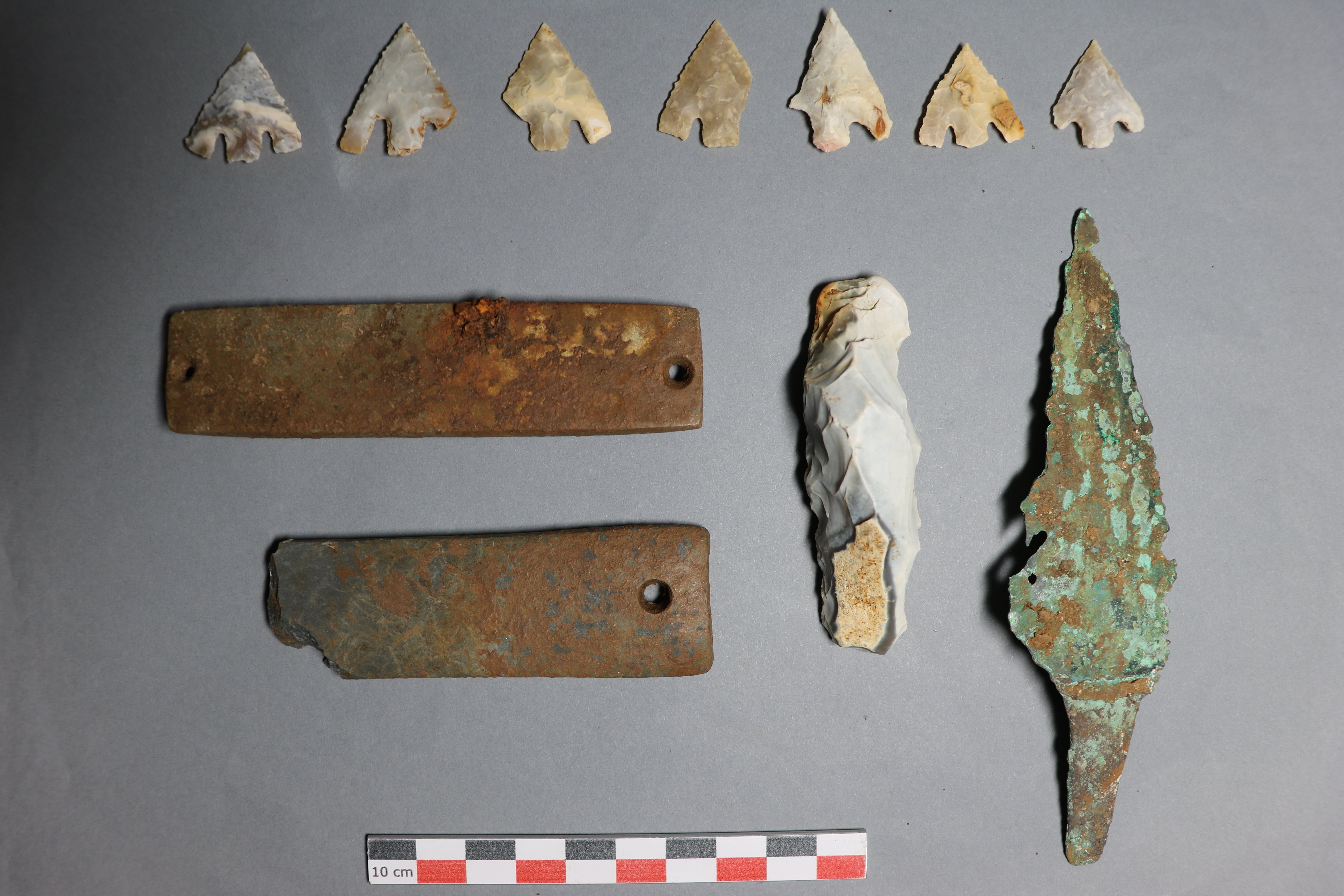 seven flint arrowheads laid out above two archer's braces, a flint lighter, and a copper alloy dagger on a grey table