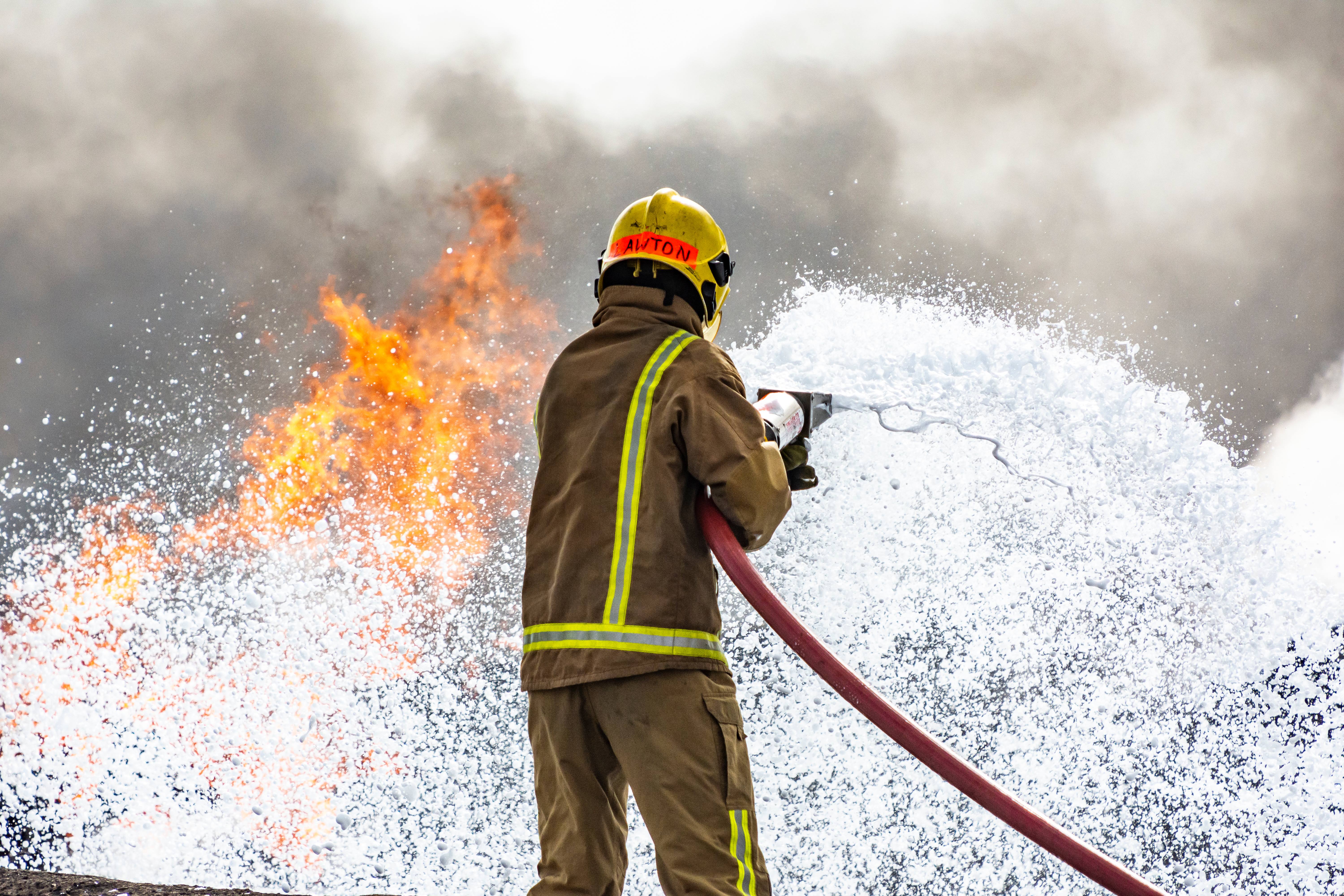 A firefighter stands with their back to the camera. They are spraying a foam onto a fire which cascades from a red hose. 