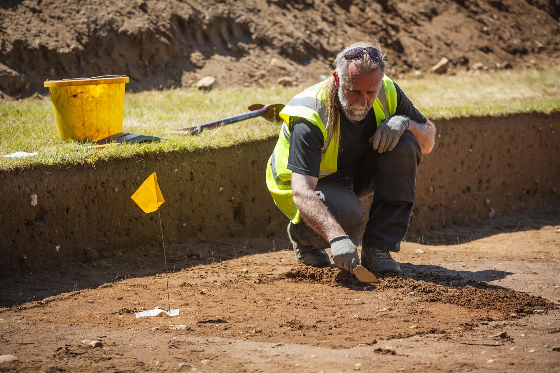 A photo of a older middle aged man in a high visibility jacket and with his sunglasses on his head is kneeling down in an excavation ditch and is examining the soil. There is a small yellow flag on a long stick to his right and a large yellow bucket and space is behind him on the grass. 