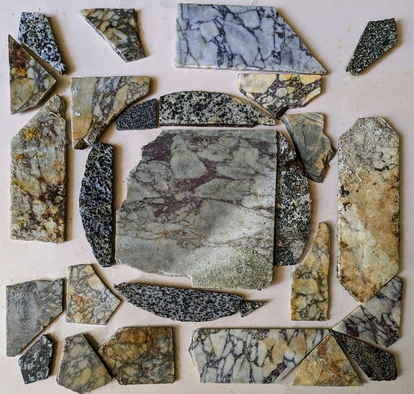 Multiple pieces of stone of different patterns and type have been arranged on a table to show how the mosaic fitted together. At the centre is a larger square stone surrounded by pieces with a rounded exterior edge. Around these are shards of various shapes and lengths. 