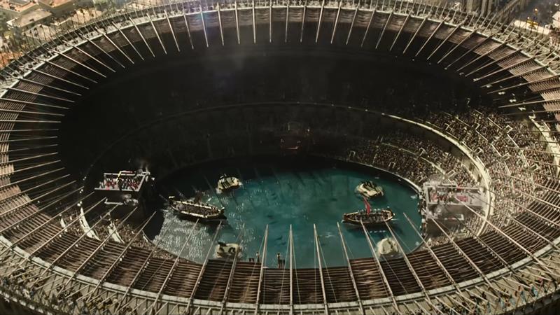 A still image from the Gladiator II trailer. The image shows a bird's eye view of the Colosseum filled with water. There are a few boats in the water and the dark forms of sharks swimming within. 