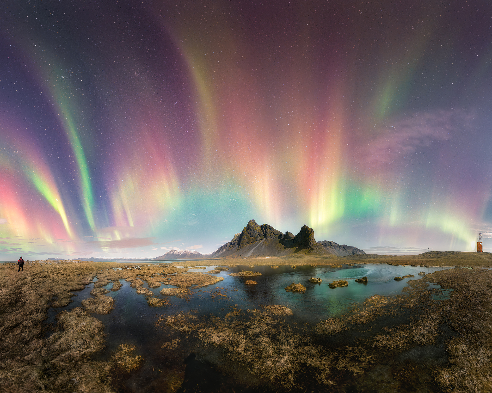 A distant mount and above aurorae of multiple colors. The photographer stands to the left of the photograph and in front there is a little lake