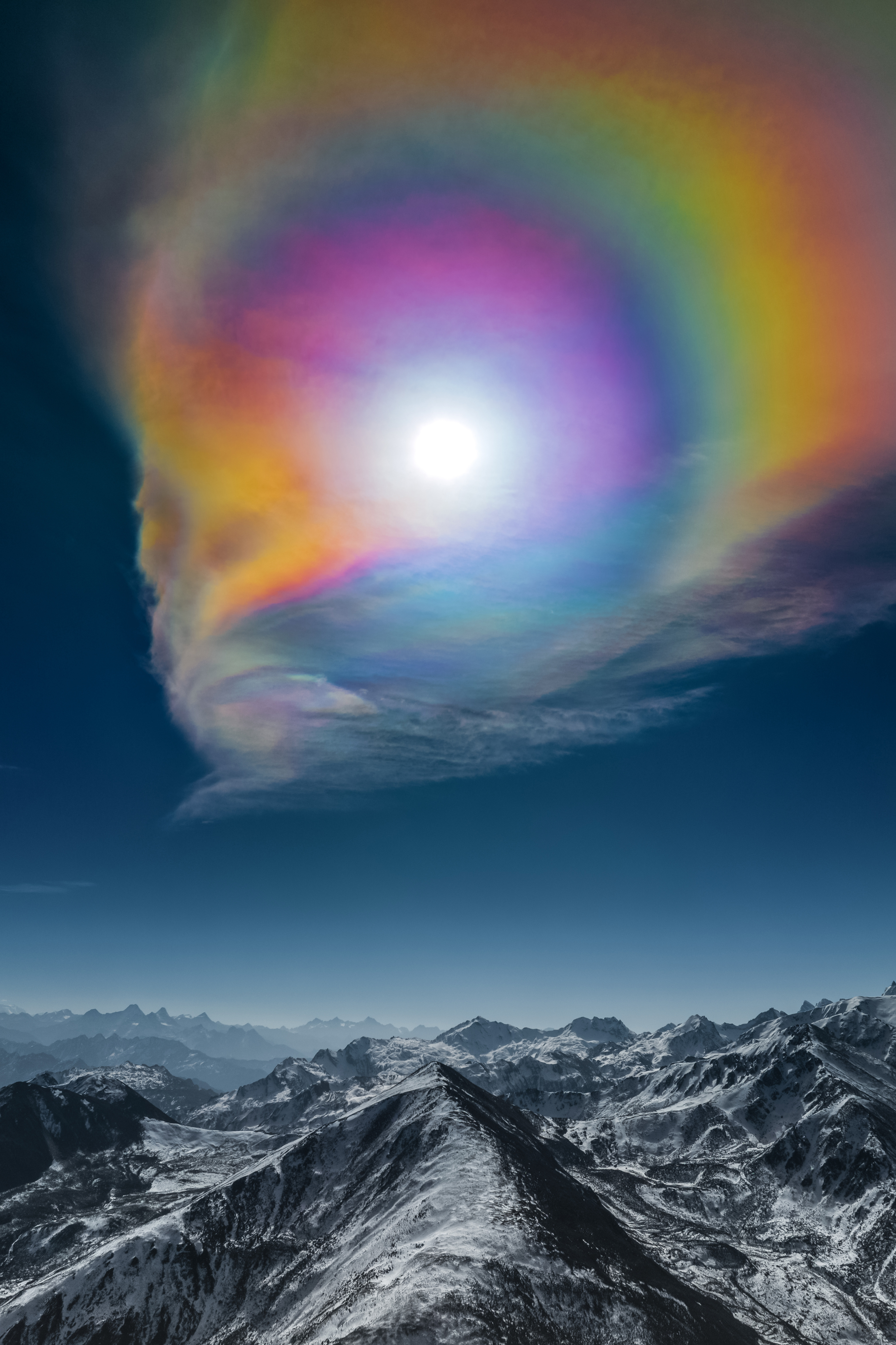 A colorful circular structure is seen withing the clouds surronding the sun. Below the snowy peaks of the himalayas