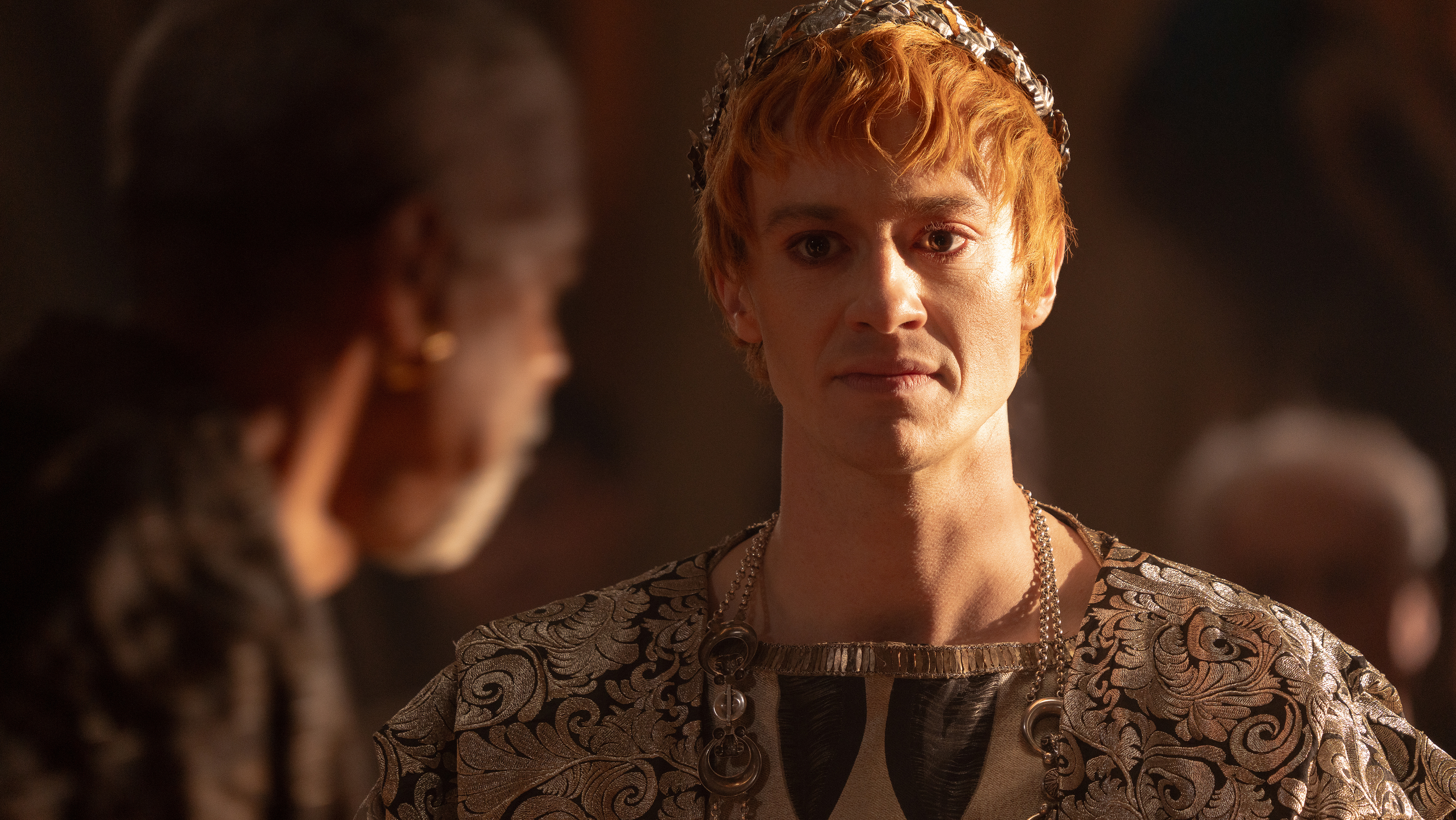 A close up of Joseph Quinn as Geta. He is wearing silver embroidered robes and a metal laurel leaf crown. His skin is overly pale and his hair is a light ginger tone.