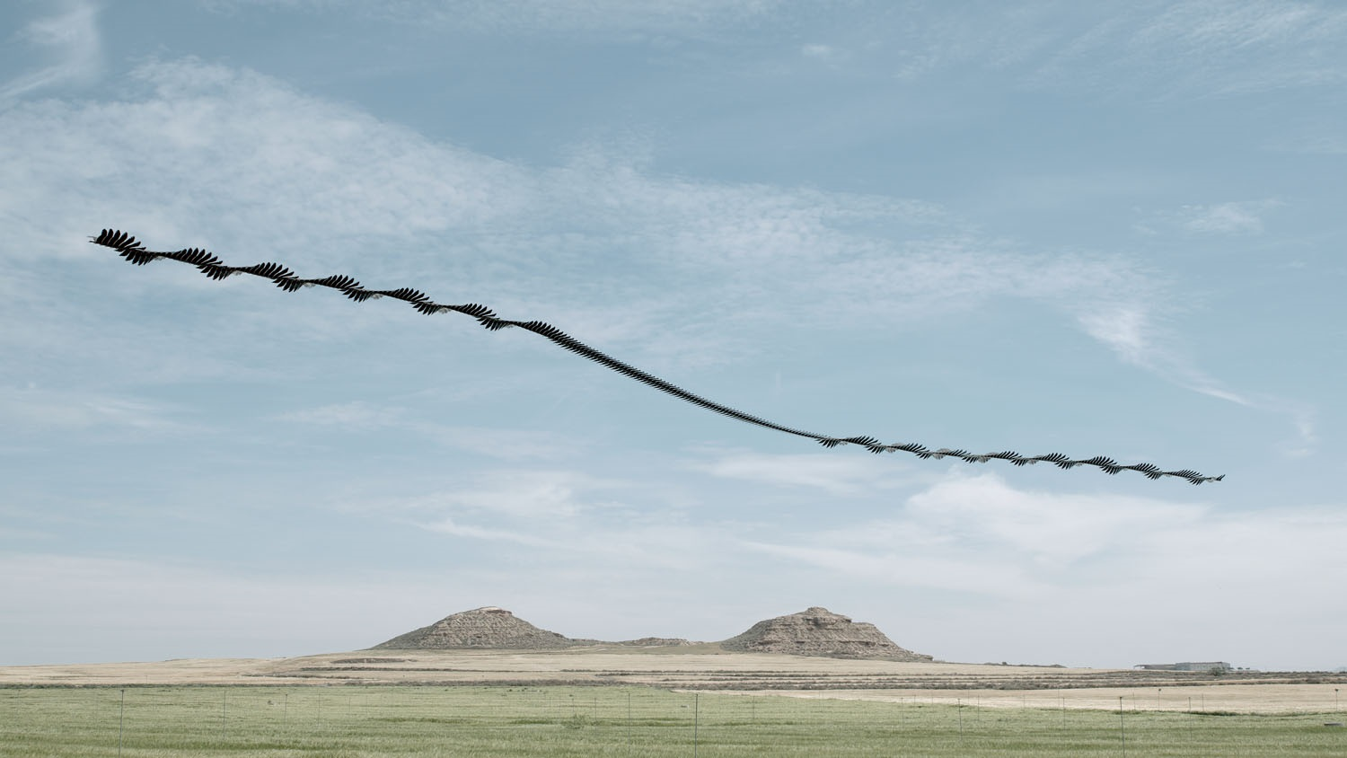 lanscape with grassy scrub and two rocky hills in the distance with large expanse of pale slightly cloudy sky, overlaid with dark black helical lines to show the path of a red kite.