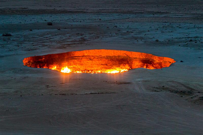 A photo of Darvaza crater taken from a distance. The shot shows the hole large crater from an elevated position in the open desert. The photo was taken after sunset so the whole hole appears to glow with flames. 