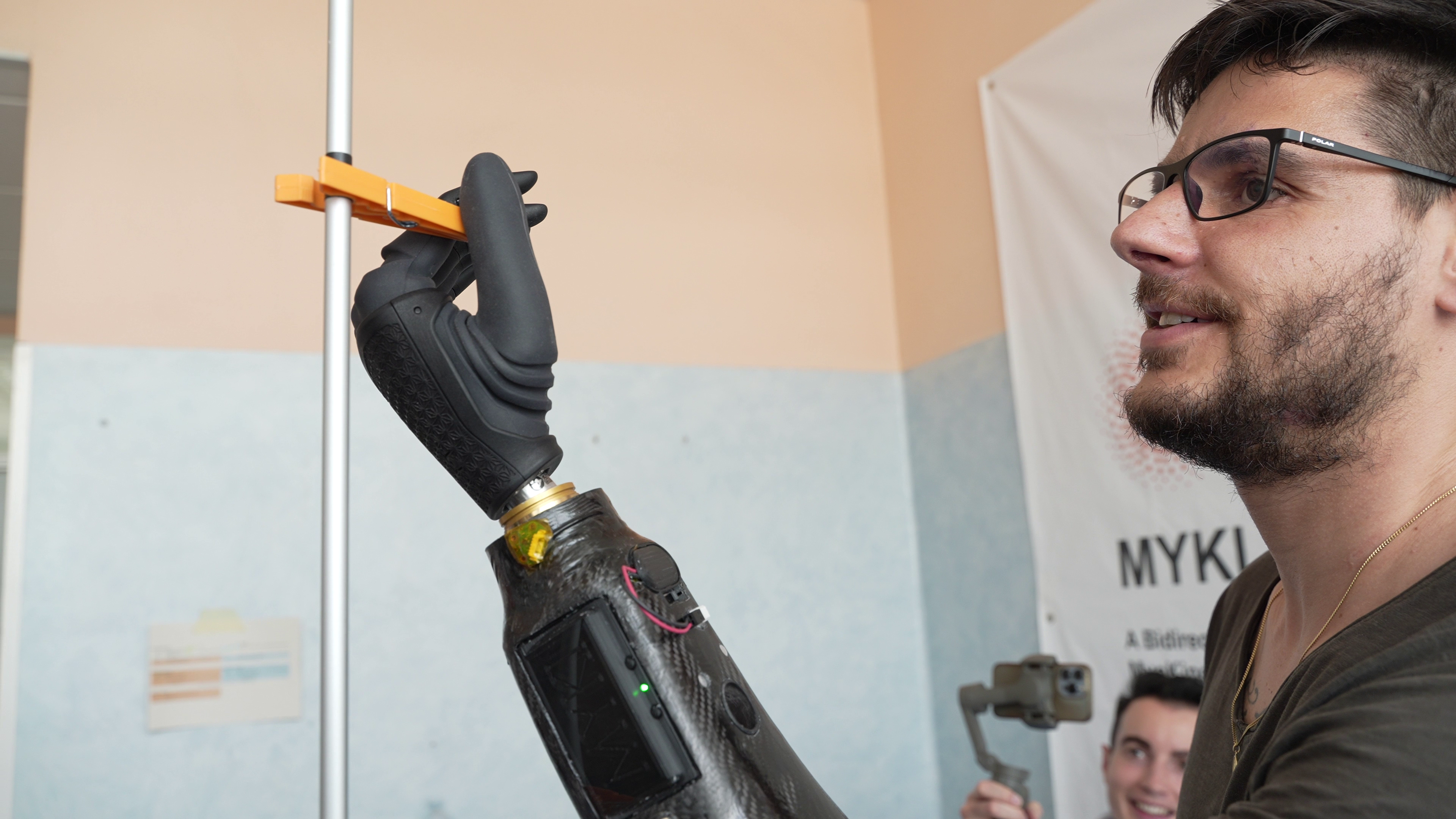 man wearing glasses operates orange clothespin with a robotic prosthetic hand