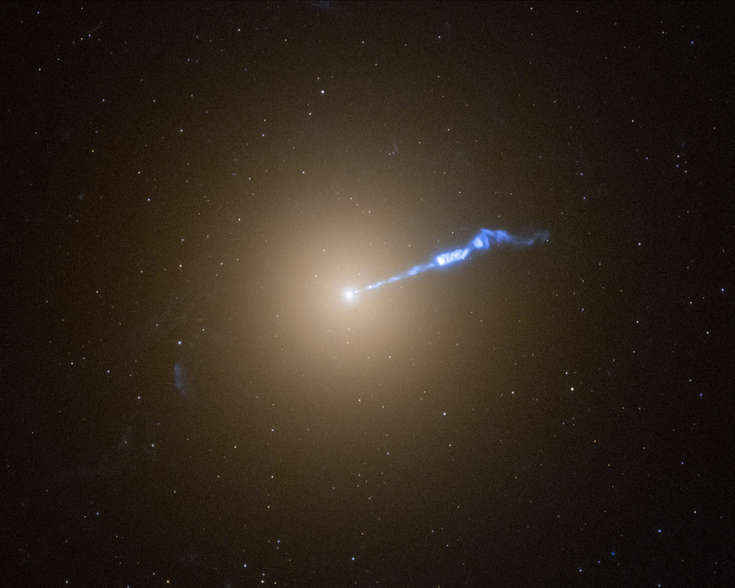 A Hubble photo of galaxy M87, which resembles a translucent, fuzzy white cotton ball. The brightness decreases gradually out in all directions from a bright white point of light at the centre. A wavy blue-white jet of material extends from the point-like core outward to the upper right, about halfway across the galaxy. Stars speckle the background