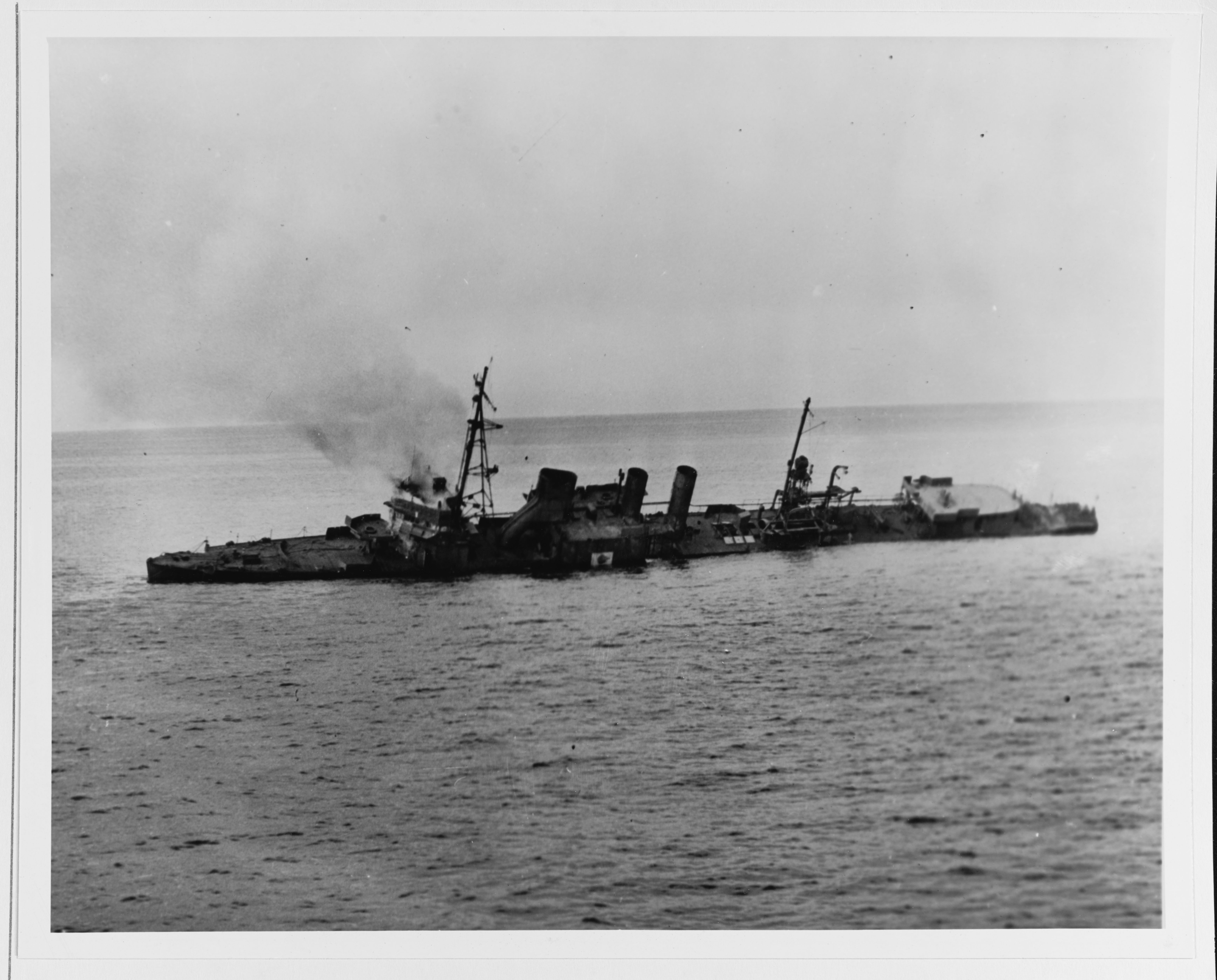 A black and white photo showing the USS Stewart as it was sinking below the water. The ship has tipped towards the camera on its left side and there is a plume of smoke rising from near the bow. 