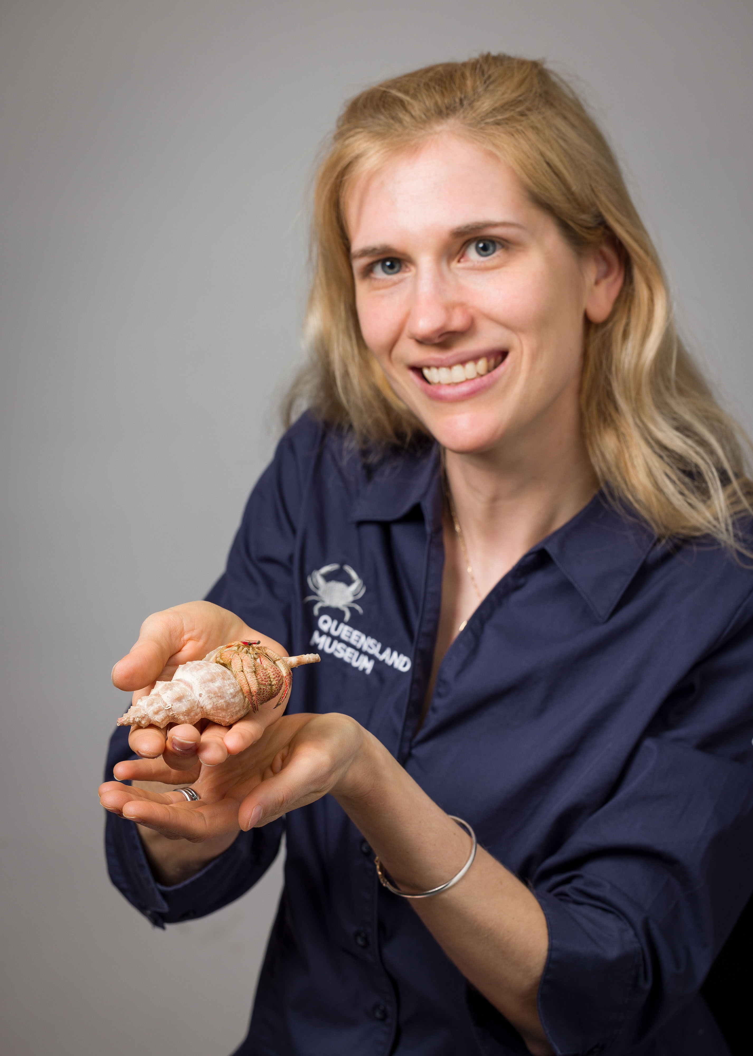 Dr Marissa McNamara, a white woman with blonde hair wearing a dark blue Queensland Museum shirt, smiles at the camera while she holds the Strawberry Claws crab