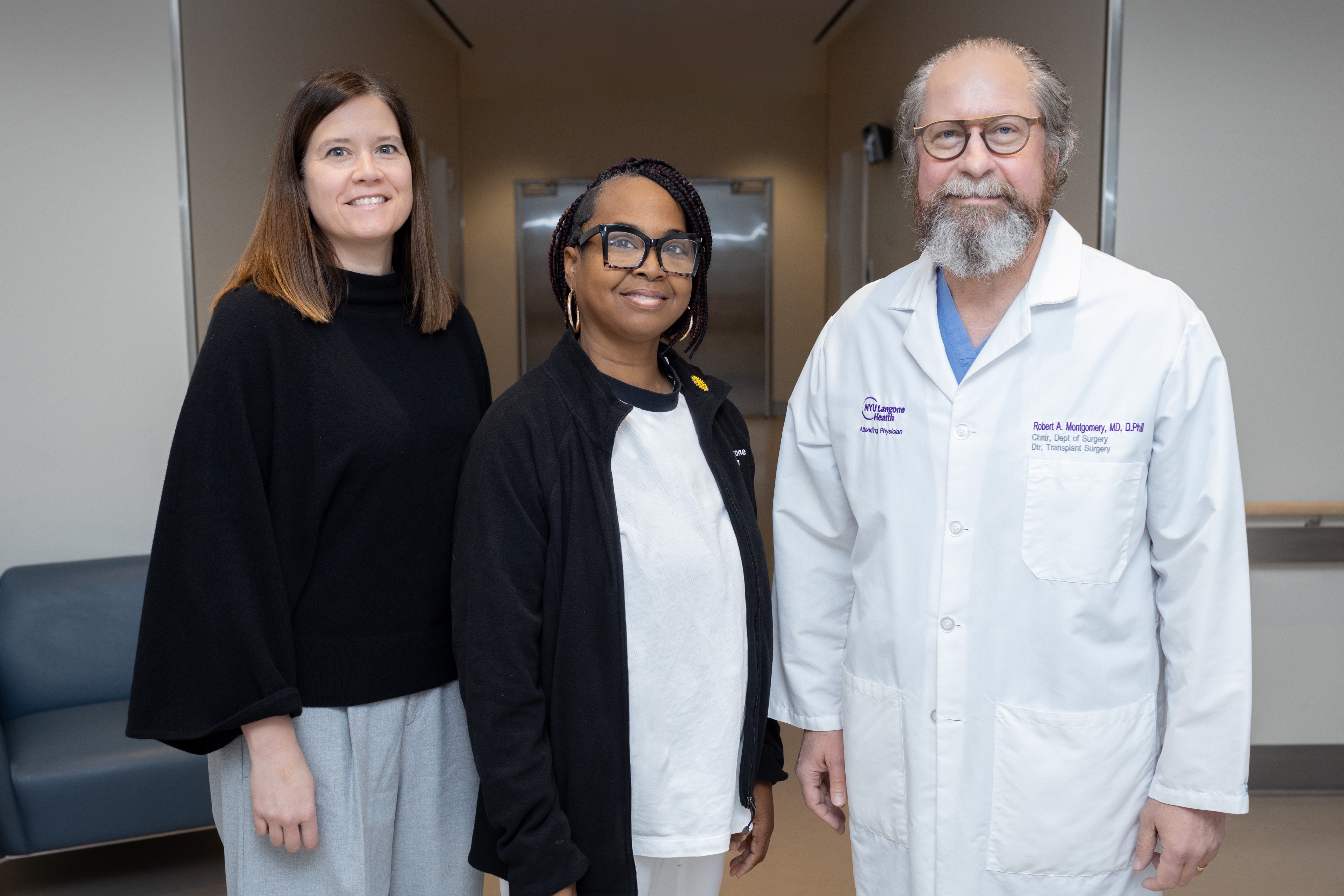 three people standing together and facing the camera smiling; a white woman with shoulder length brown hair wearing a black top and grey trousers, a black woman with braided hair wearing large spectacles, a black jacket, and white top and trousers, and a white man with gray hair and beard, wearing round spectacles and a white coat