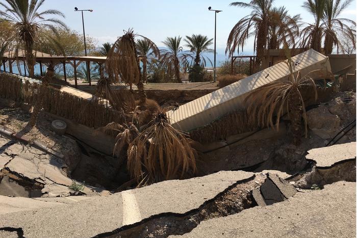 A sinkhole in the middle of a road. The ground has collapsed in and the debris includes what appear to be a bus and a lory. There are palm trees growing in the middle of fissure. In the background, the lake is visible against a blue sky. 