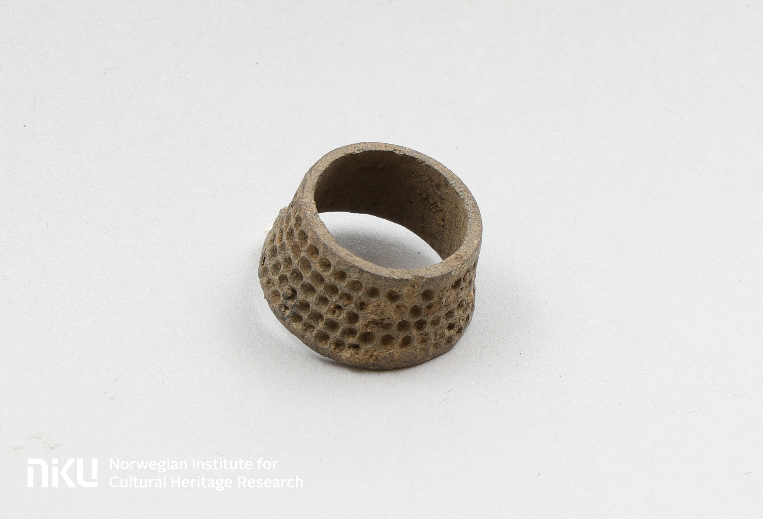 A photo of the thimble. It looks like a pot with not top or bottom. It has rows of small shallow holes running along its sides.