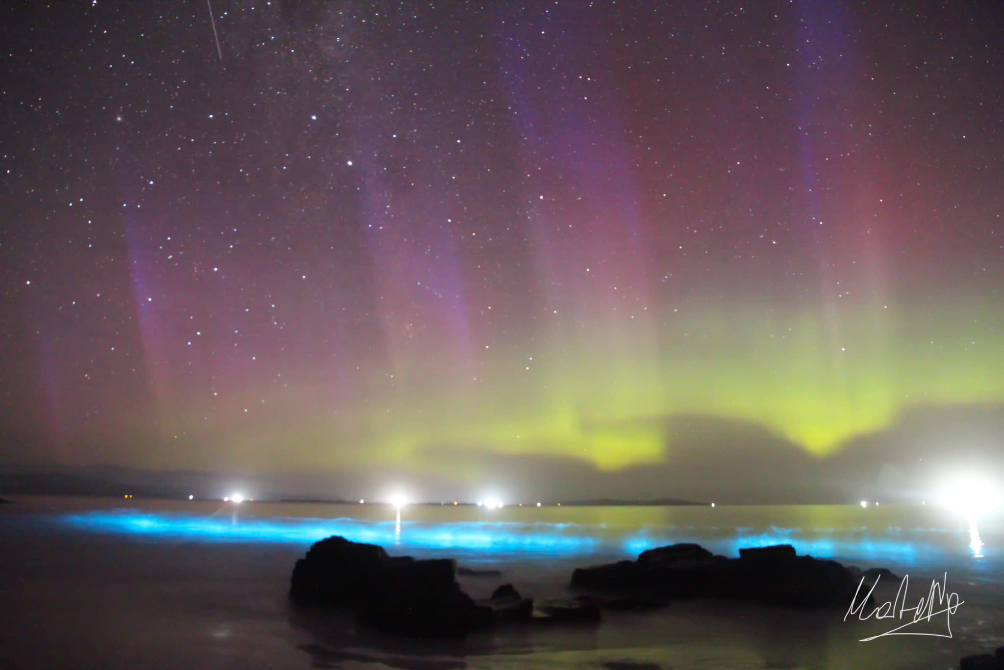 Some auroras come with beams, captured here with bioluminescence breaking on the beach.