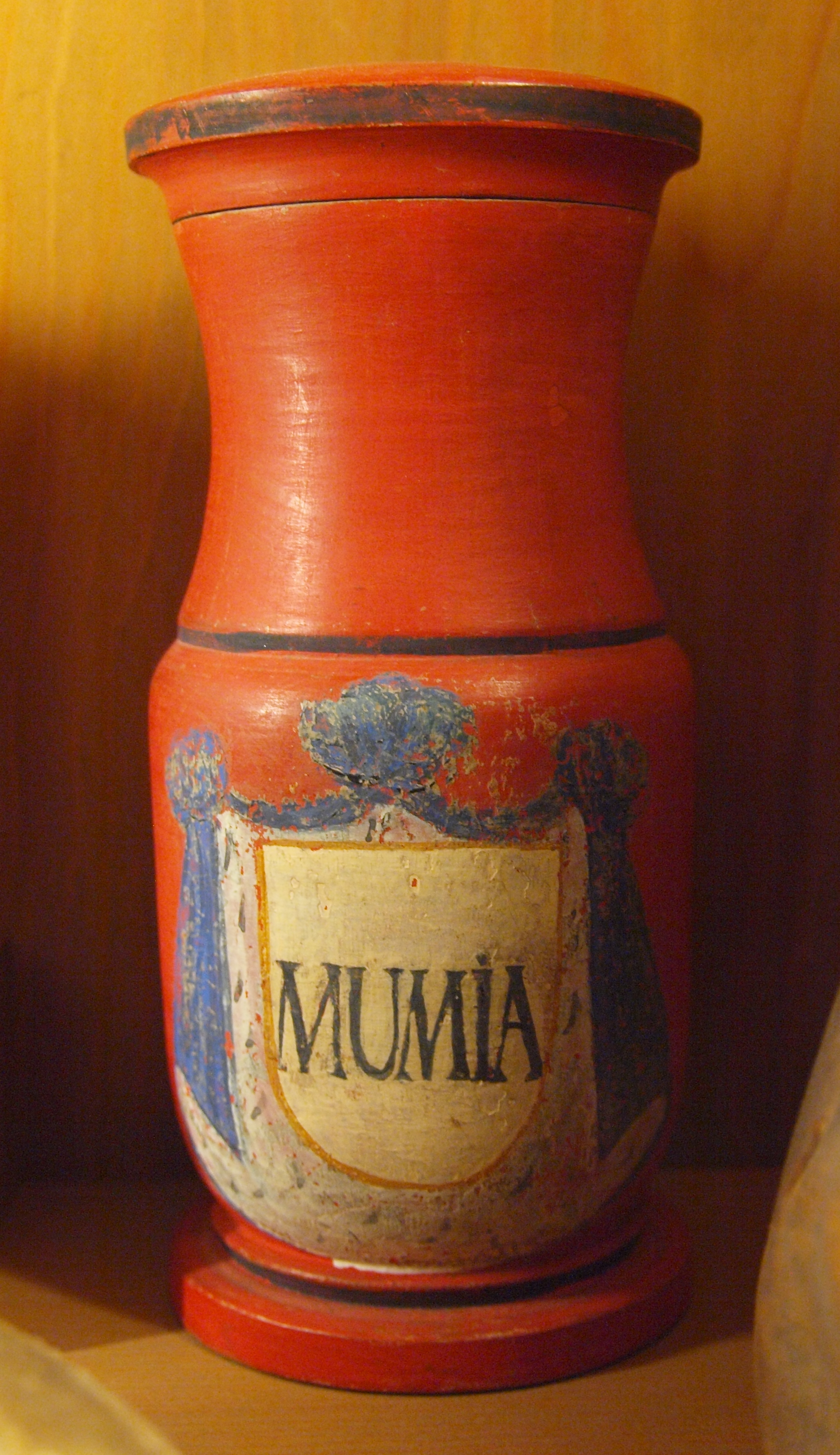 A jar shaped like an urn painted in a deep orange/red colour. It has a blue rim and two blue lines at the base of its neck and at the base of its body. The word 