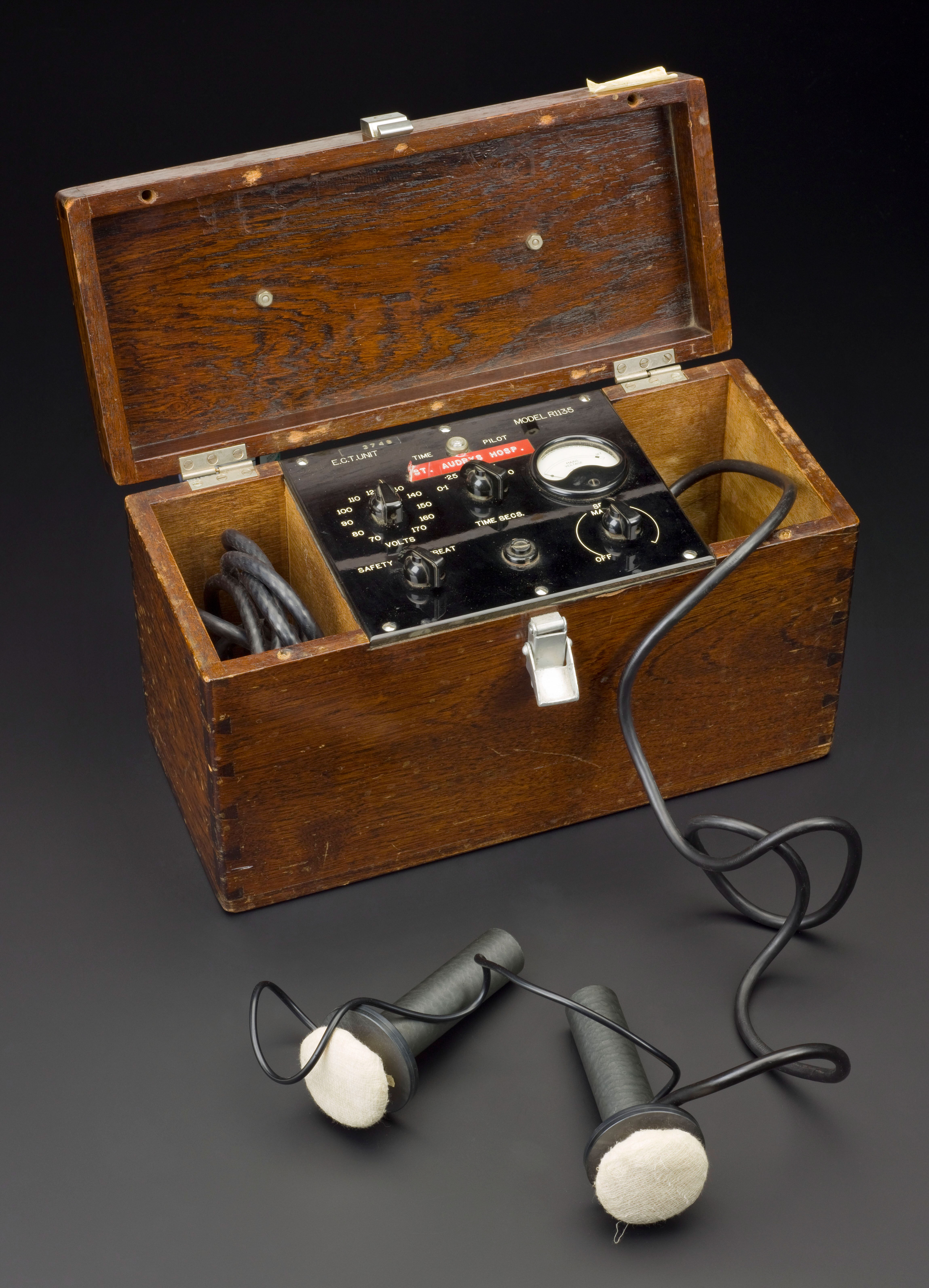 A photo showing a wooden box with an open lid. Inside the box is a panel with various dials and buttons. Either side of the panel is space for cables to be folded up. The right hand side space has a cable extending out of the box which terminates in two small microphone-like shaped objects with white ends. e