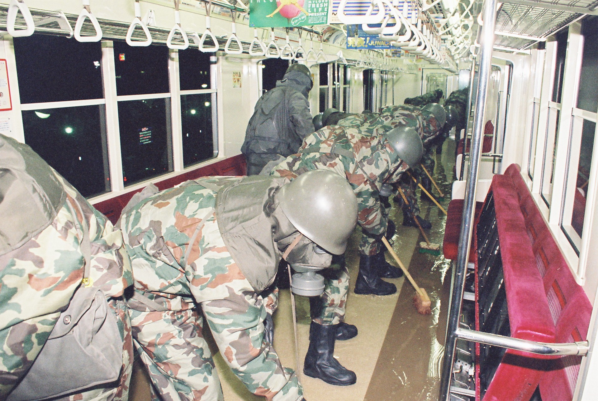 A line of military officers are scrubbing the floor of a train in the Tokyo underground. The men are wearing camouflaged suits, gas masks and helmets. They are all leaning over brooms and rubbing a liquid that covers the floor of the carriage.  