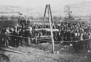 A photo showing the supposed Cardiff giant being lifted from the ground. The photo is black and white and grainy. It shows a dozens of people gathered around a scaffold that appears to be raising the giant out of the ground. The giant is laying horizonal in the middle of the shot and the foreground shows open grass and then a rope fence in front of the gathered people.  