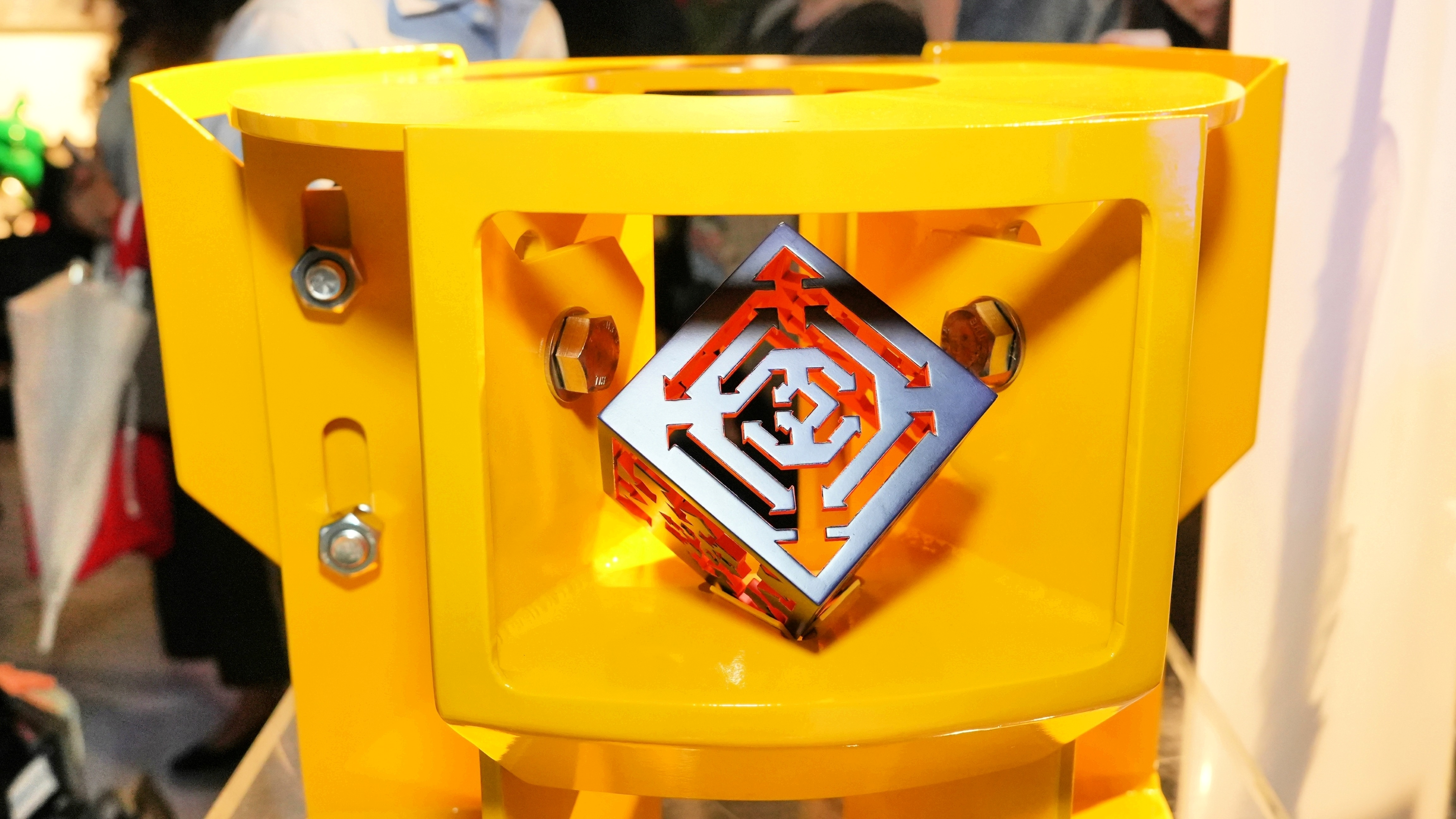 bright yellow container with a metal cube inside; the face pointed towards the camera has a pattern of geometric arrows cut out of the surface