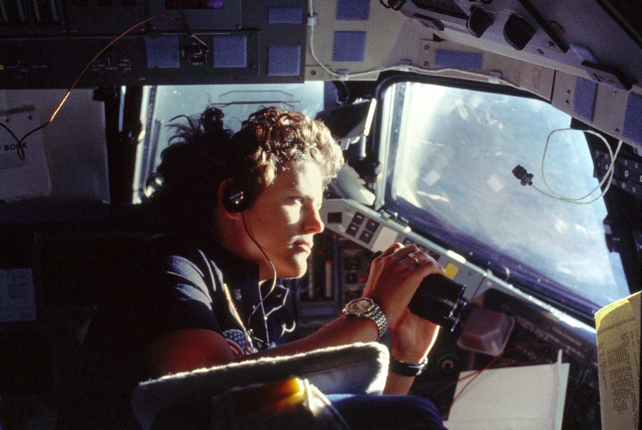 Sullivan is floating in the cockpit of shuttle holding a binocular. The Earth is visible in the window