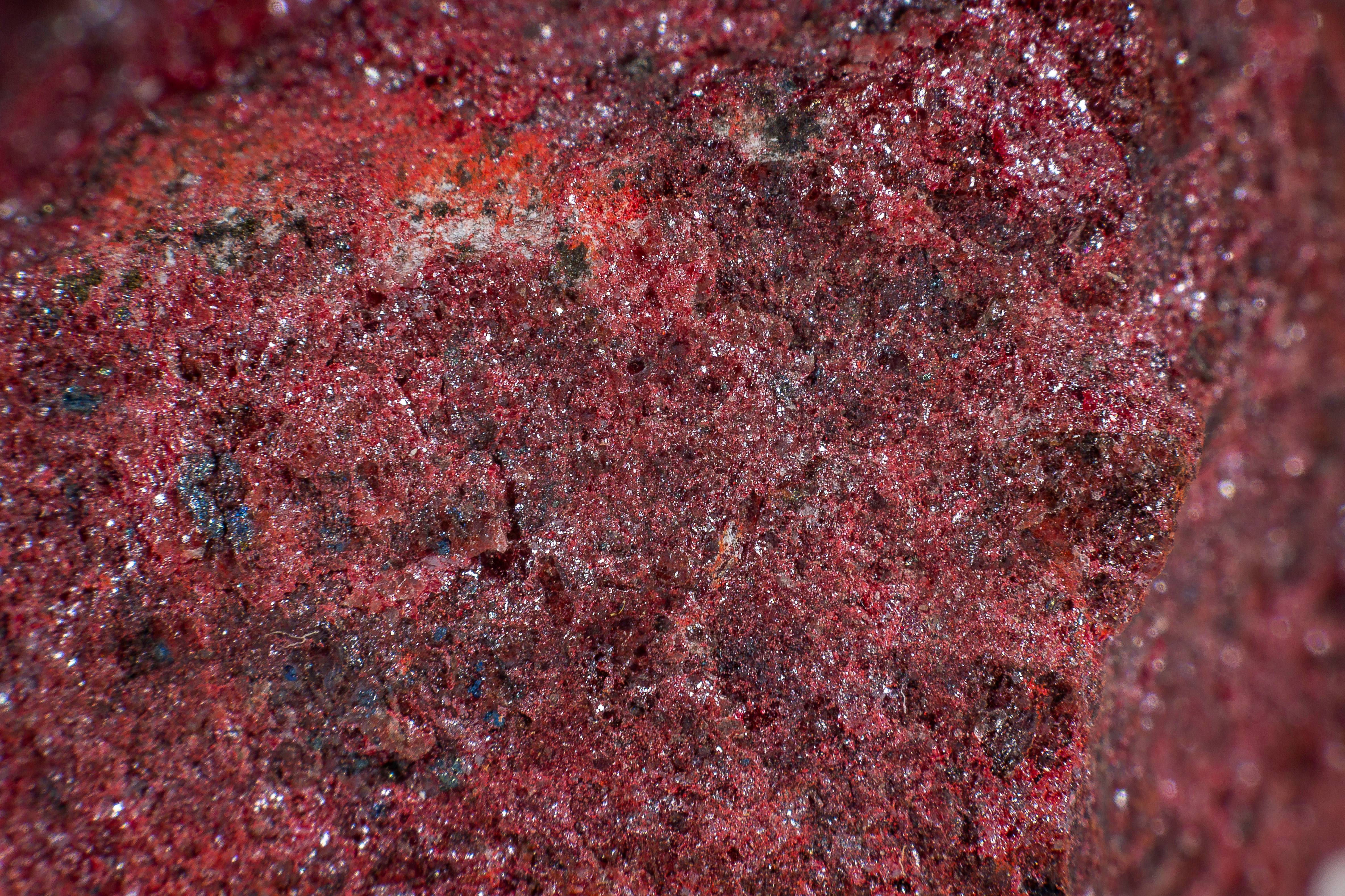 A close up of cinnabar mineral stone. The surface is a deep read with a rough crystalline texture. Some patches of crystal are bigger than others, giving it a darker red look.  