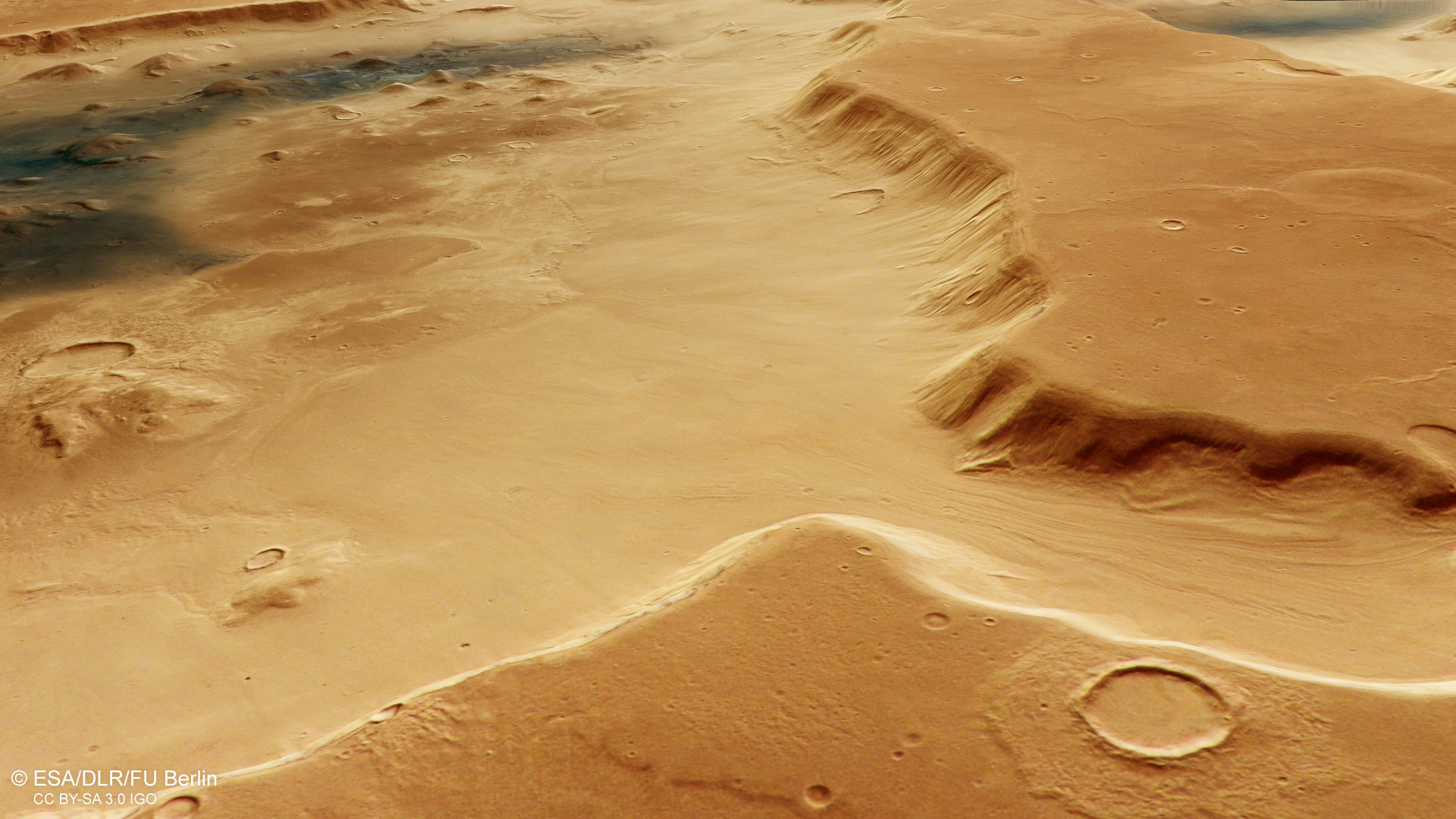 A sweeping view along the crater rim of Deuteronilus Cavus, which transects the image from bottom right to centre-top, focuses on the smooth debris flows that have slid towards the centre of the crater. At the right, a large chunk of the crater wall is missing, forming a U-shaped valley with a grooved floor. Towards the centre of the crater – the far top left of the image – a patch of dark volcanic dust covers the surface. Jumbled blocks are seen in the centre, contrasting the smoother flows around the inner walls