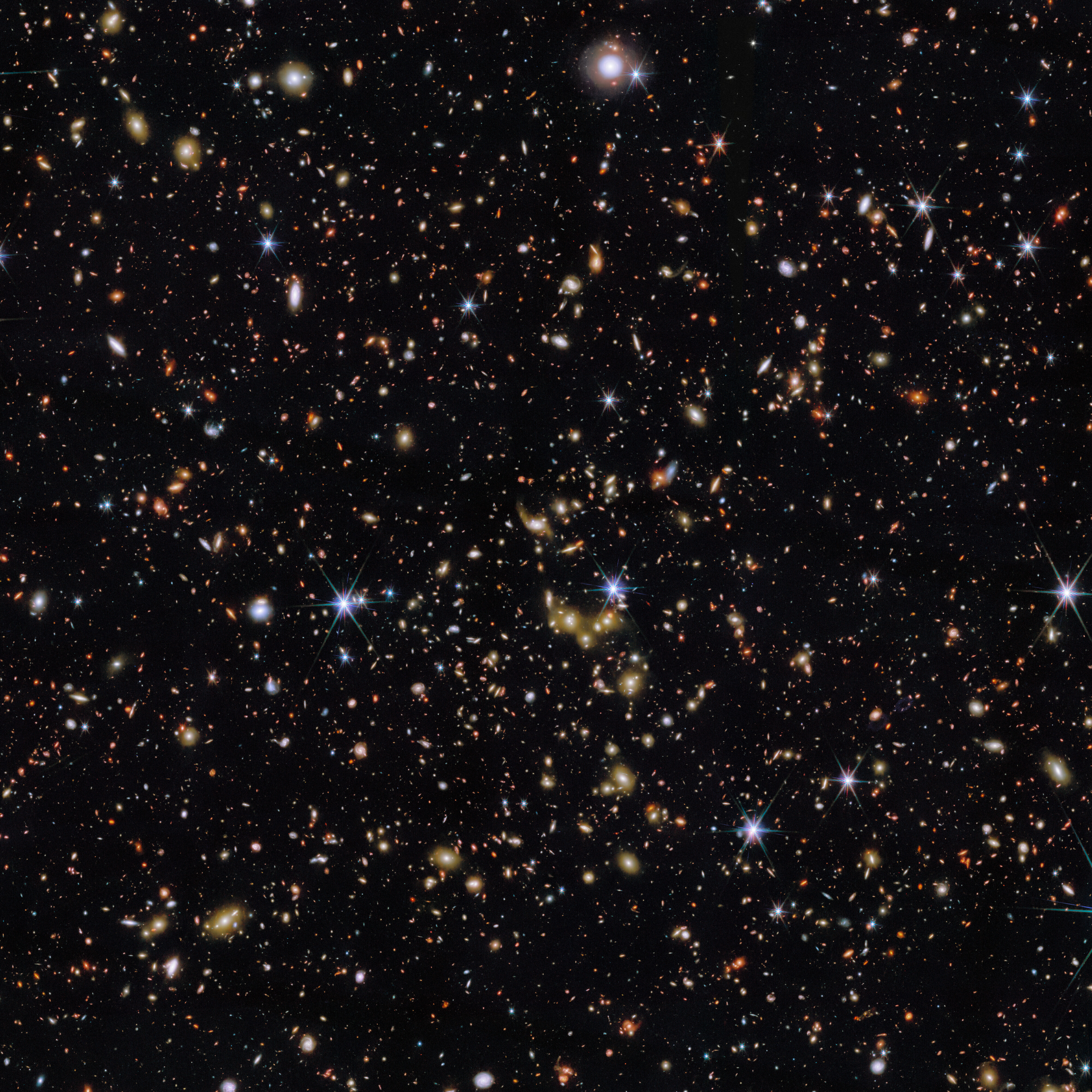 An area of deep space with thousands of galaxies in various shapes and sizes on a black background. Most are circles or ovals, with a few spirals. More distant galaxies are redder in colour and smaller, down to being mere dots, while closer galaxies are a bit larger and white or blueish. A few gold-coloured galaxies are bunched closely together in the centre. Bright stars surrounded by spikes lie in our galaxy.