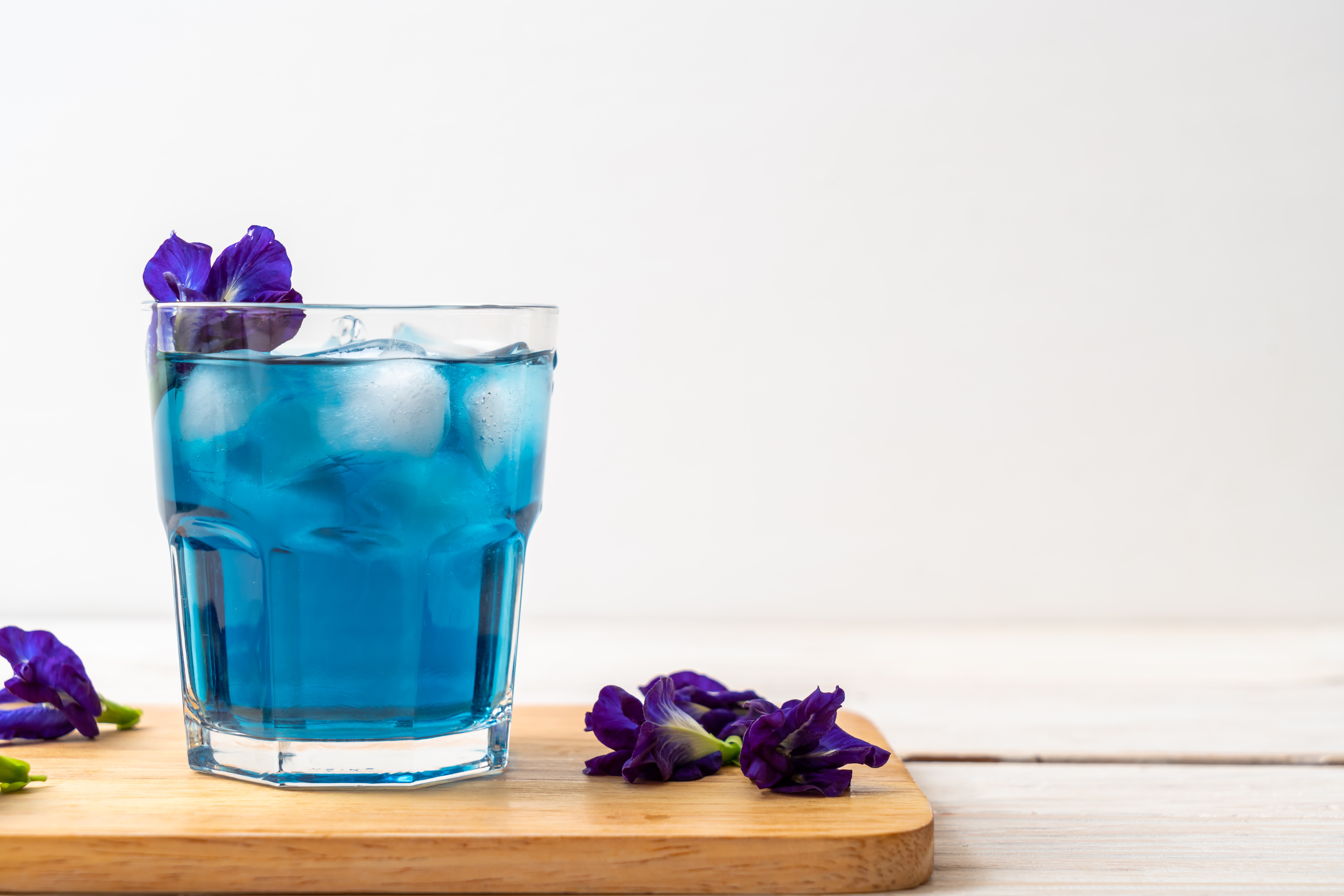 glass of blue liquid with ice; there are butterfly pea flowers on the glass and around it. It is sitting on a wooden chopping board on a light backgroun glass of blue liquid with ice; there are butterfly pea flowers on the glass and around it. It is sitting on a wooden chopping board on a light backgroun