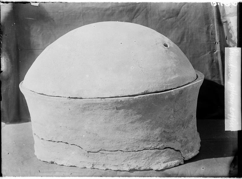 A black and white photo showing the ceramic pot the individual was buried in. The pot is pale coloured and has a rounded lid. The body is crudely made appearing lopsided towards the right. 