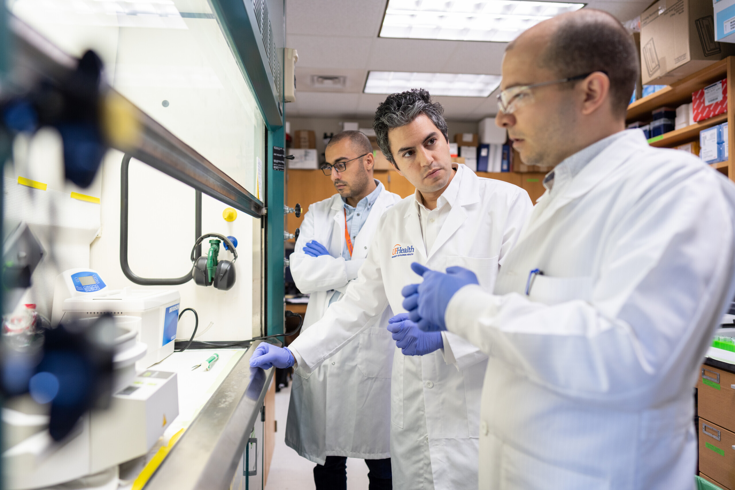 three male scientists wearing lab coats and gloves standing in front of a culture hood in a lab with lots of brown shelving units and cabinets behind them three male scientists wearing lab coats and gloves standing in front of a culture hood in a lab with lots of brown shelving units and cabinets behind them