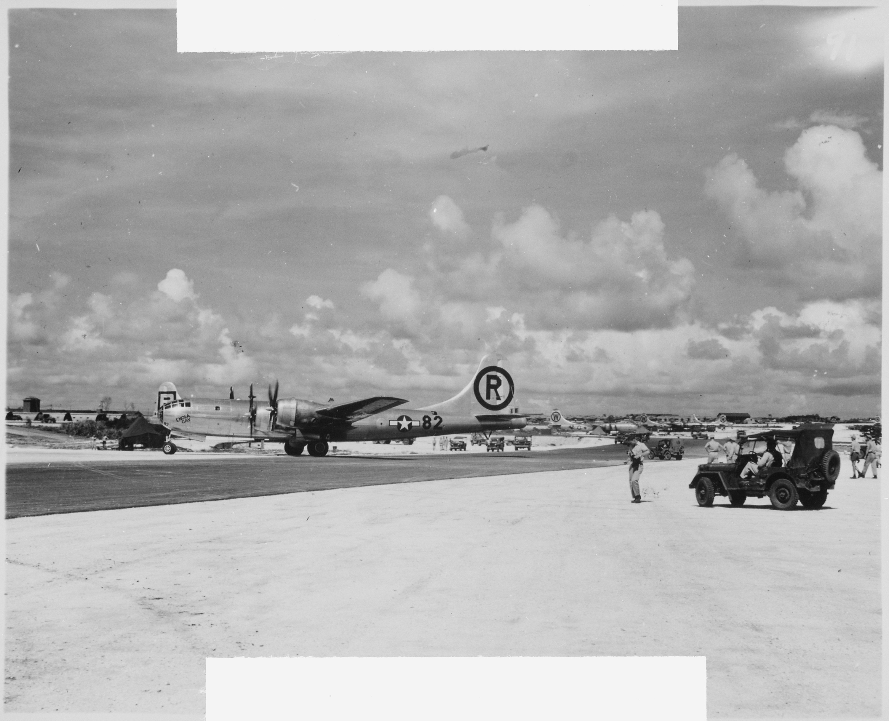 A black and white photo showing the Enola Gay on a runway. The aircraft is shown on profile, facing the left of the shot. It has twin predellas on each wings. There is a large R encircled and printed on its tail and the number 82 is printed on its tail, next to a symbol of a star in a circle with a rectangle running through it. The word 